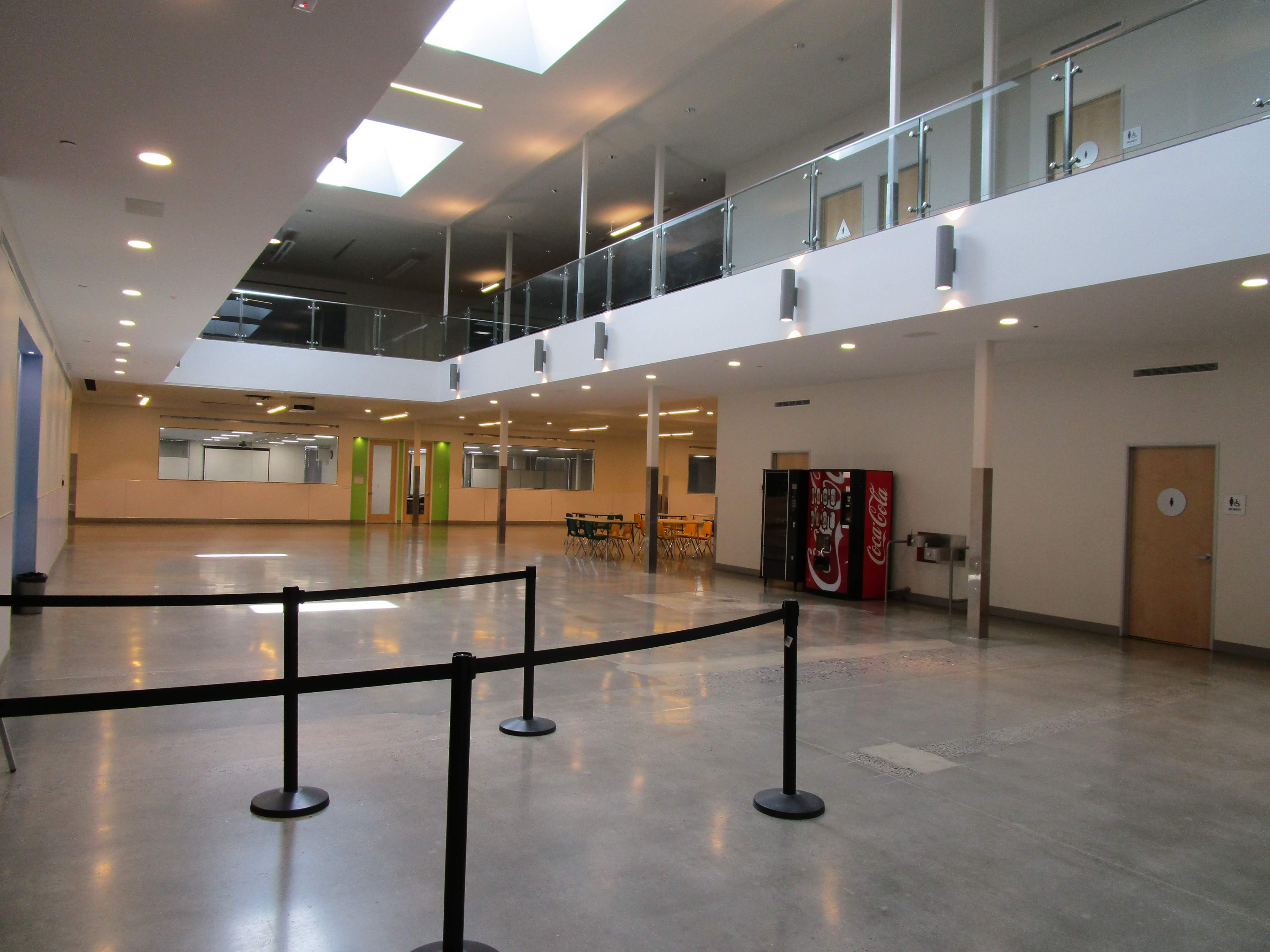 The image shows a spacious indoor area with a high ceiling, featuring a row of vending machines, tables in the distance, and a barrier with retractable cord.