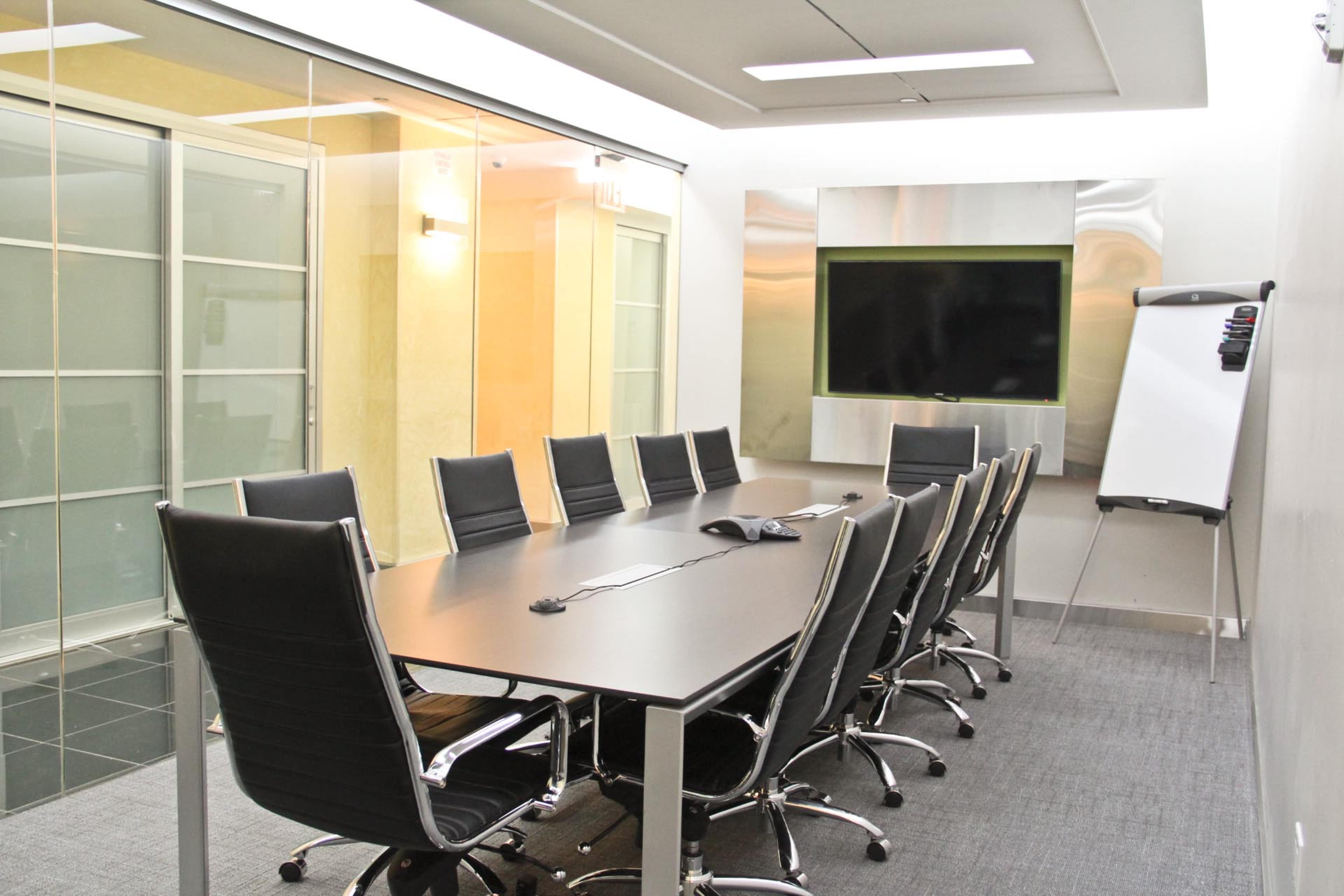 A conference room features a long table surrounded by black chairs, with a glass wall and a large screen at one end.