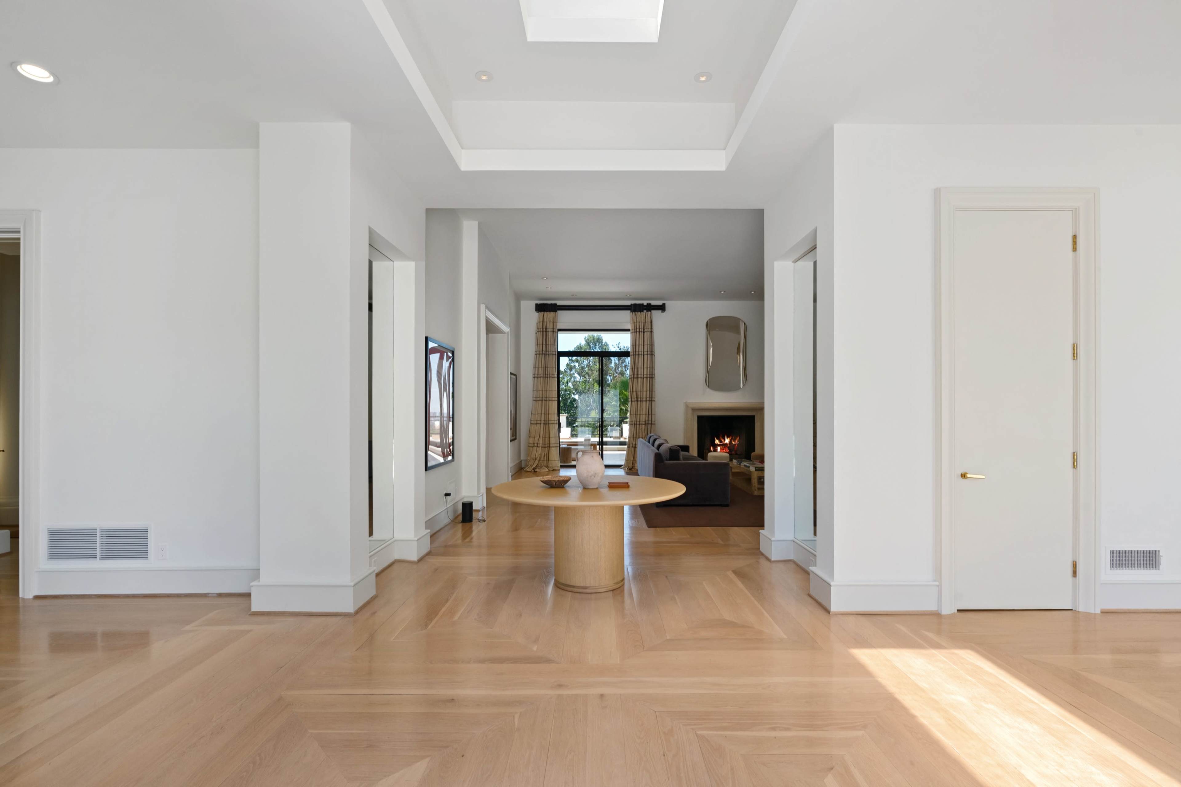 A spacious, modern interior with light hardwood floors, a round table in the center, and a view of a room with a fireplace in the background.