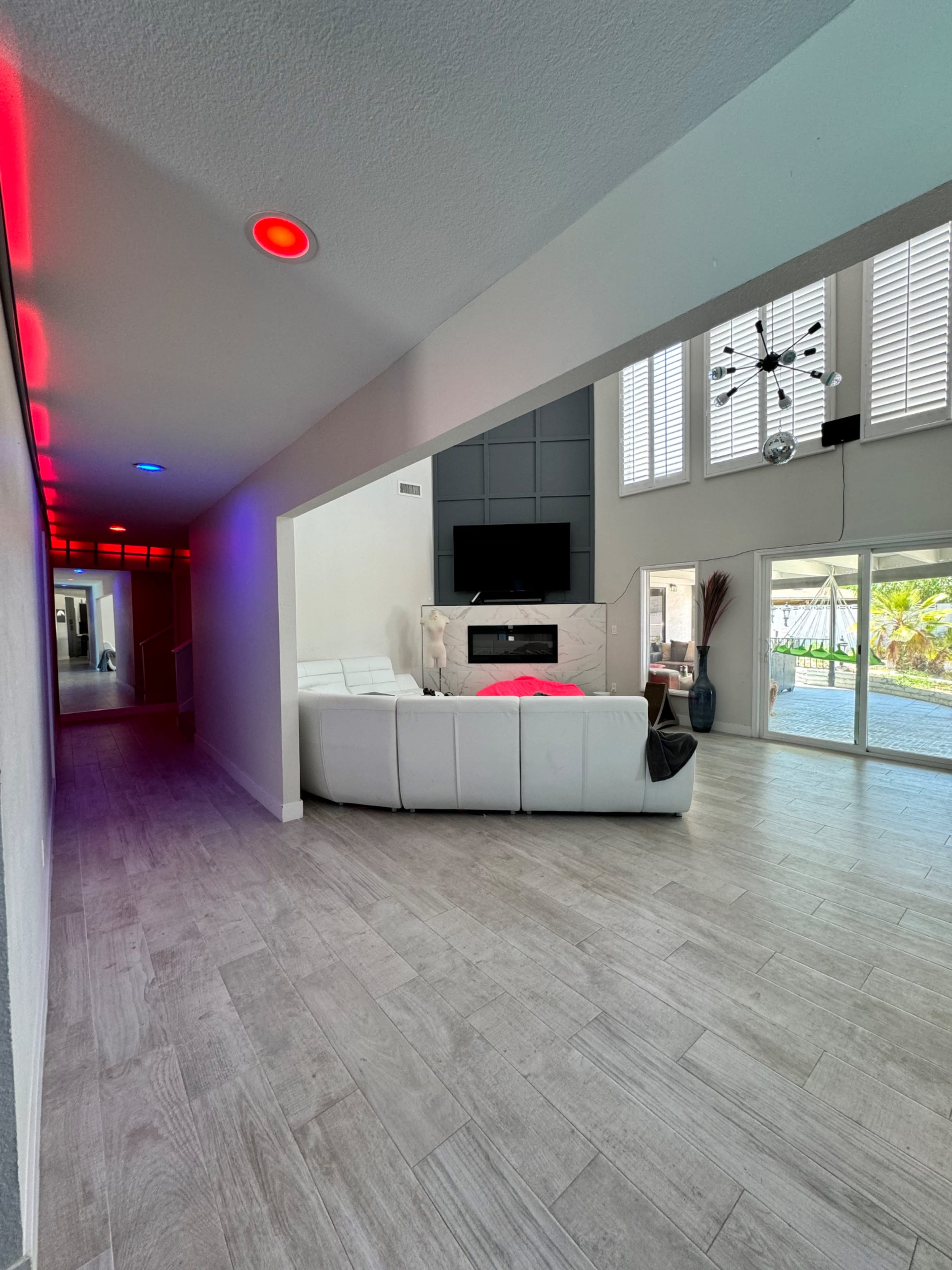 A modern living room features a sectional sofa, a wall-mounted TV, and large windows with natural light, with an adjacent corridor illuminated in red and blue lights.