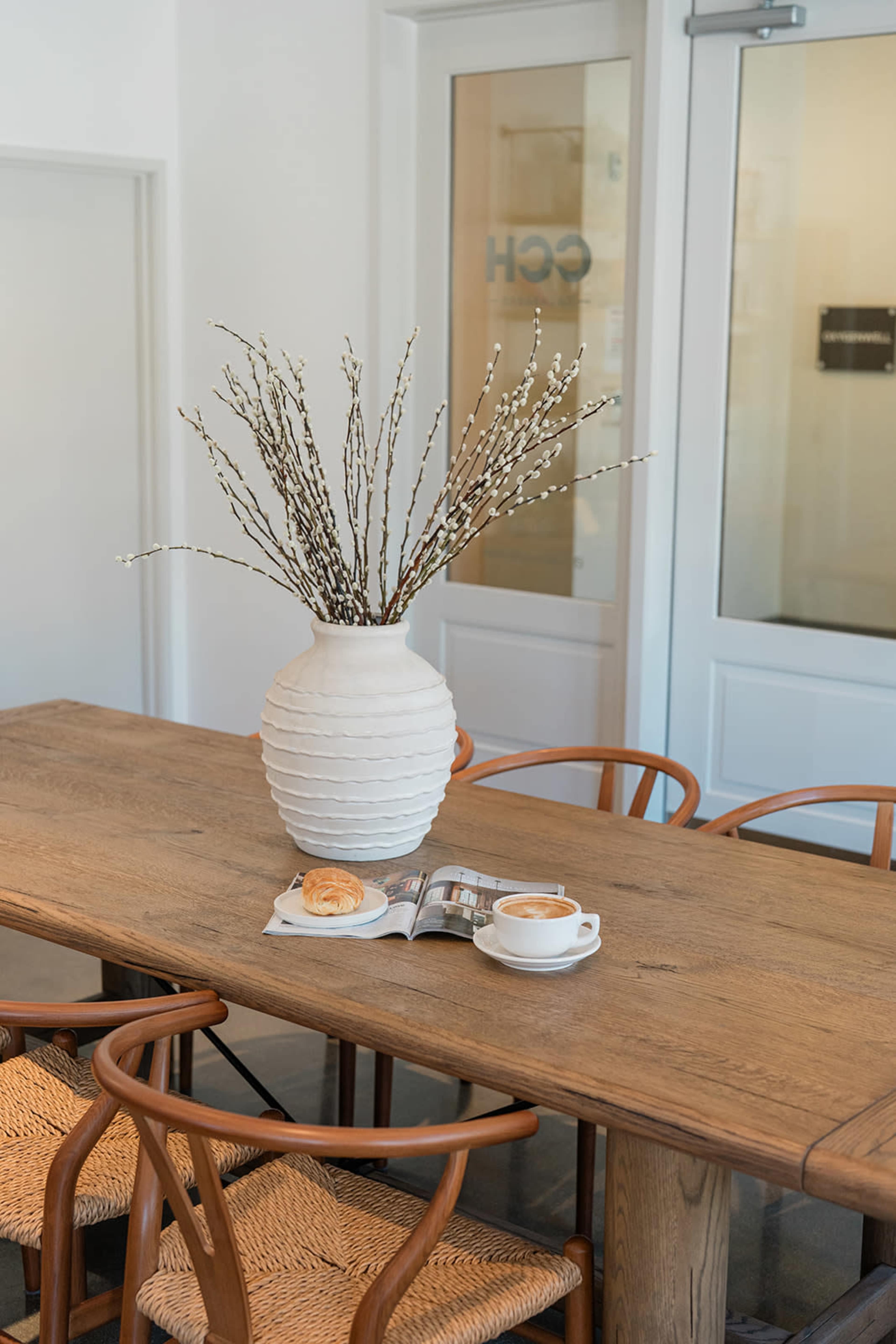 A large white vase with branches sits on a wooden table next to a cup of coffee and a pastry beside an open magazine.