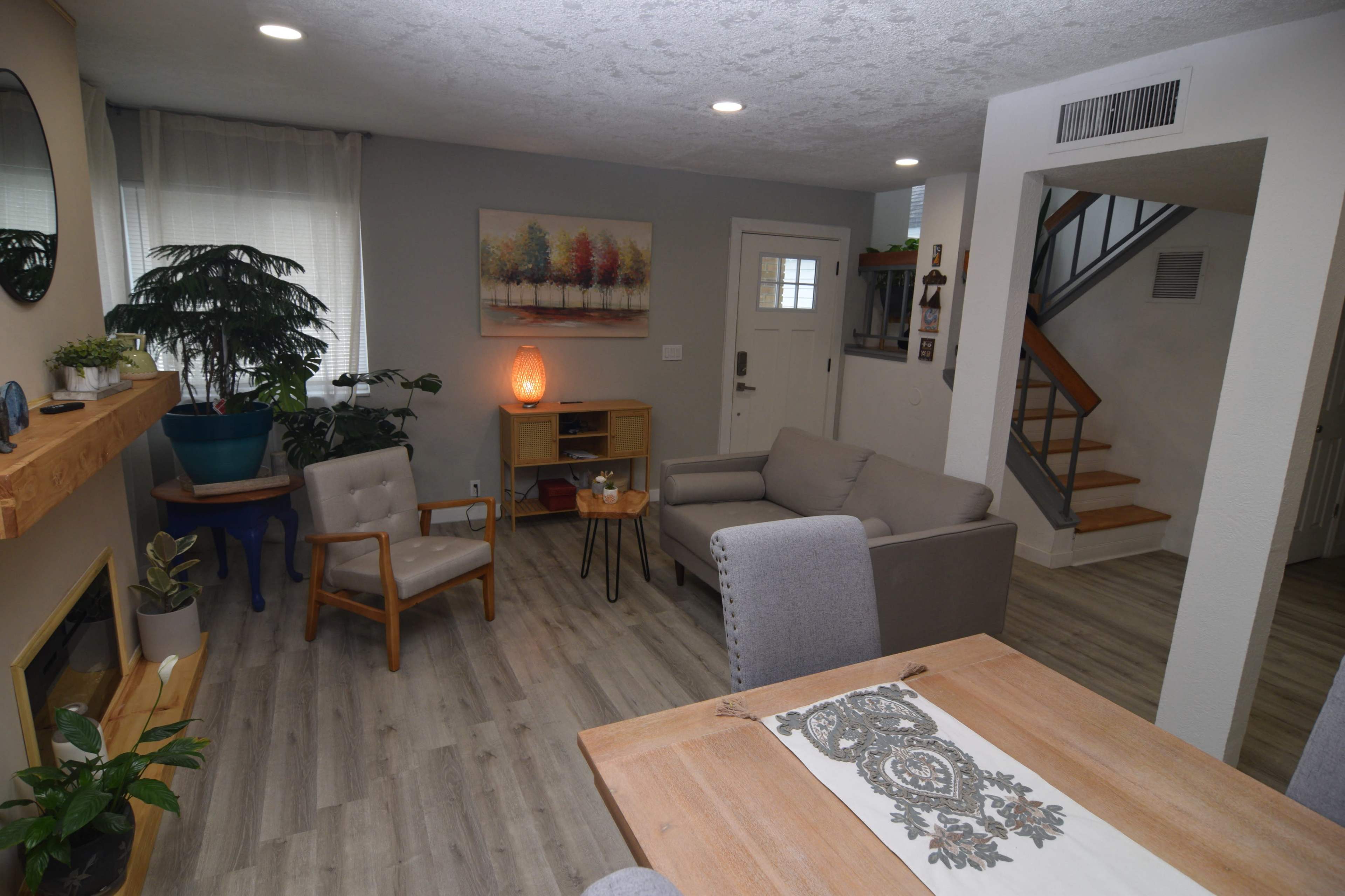 A modern living room with a comfortable seating arrangement, a wooden dining table, a small bookshelf, and a staircase in the background.