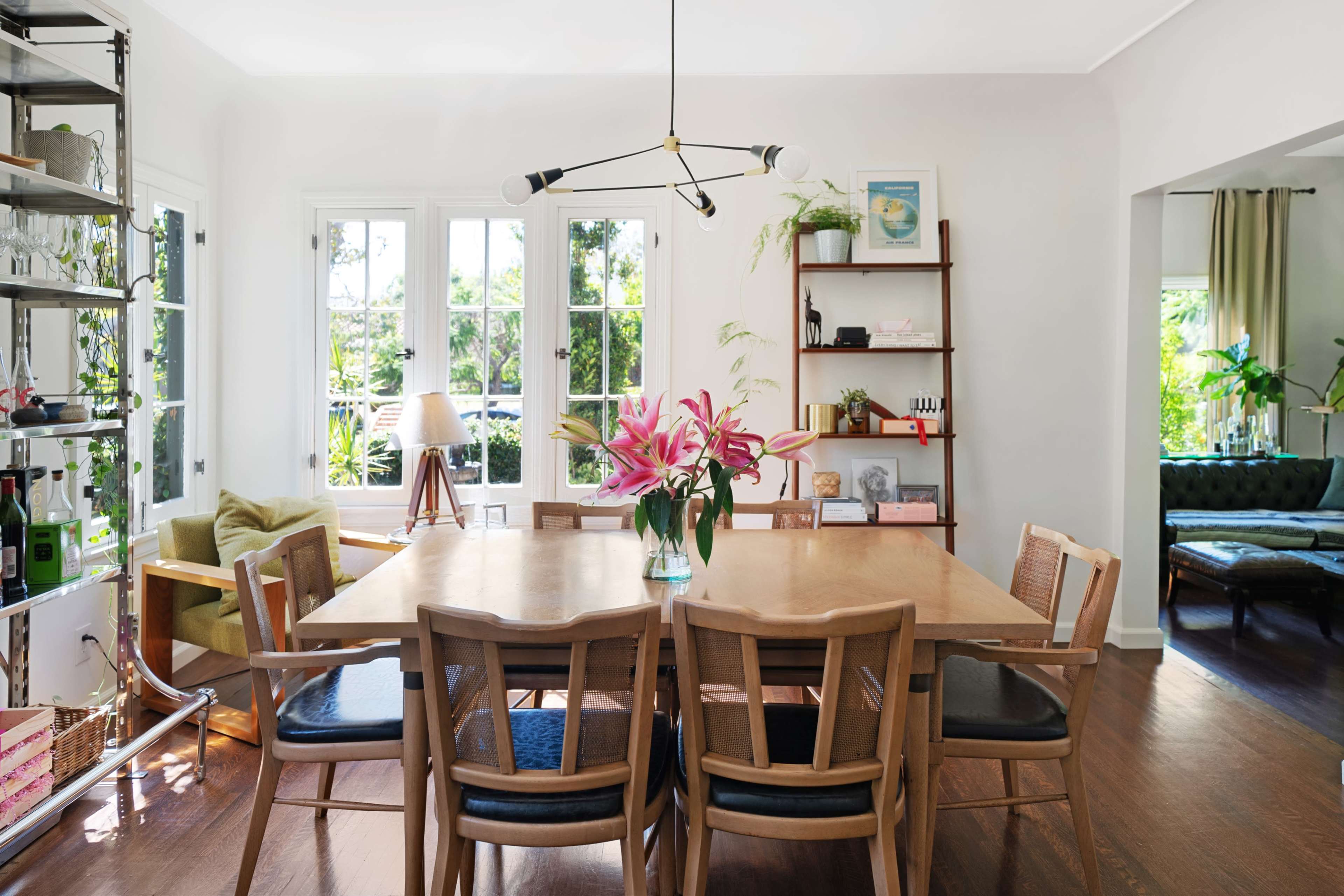 A wooden dining table surrounded by chairs is set in a bright room with large windows and greenery visible outside.