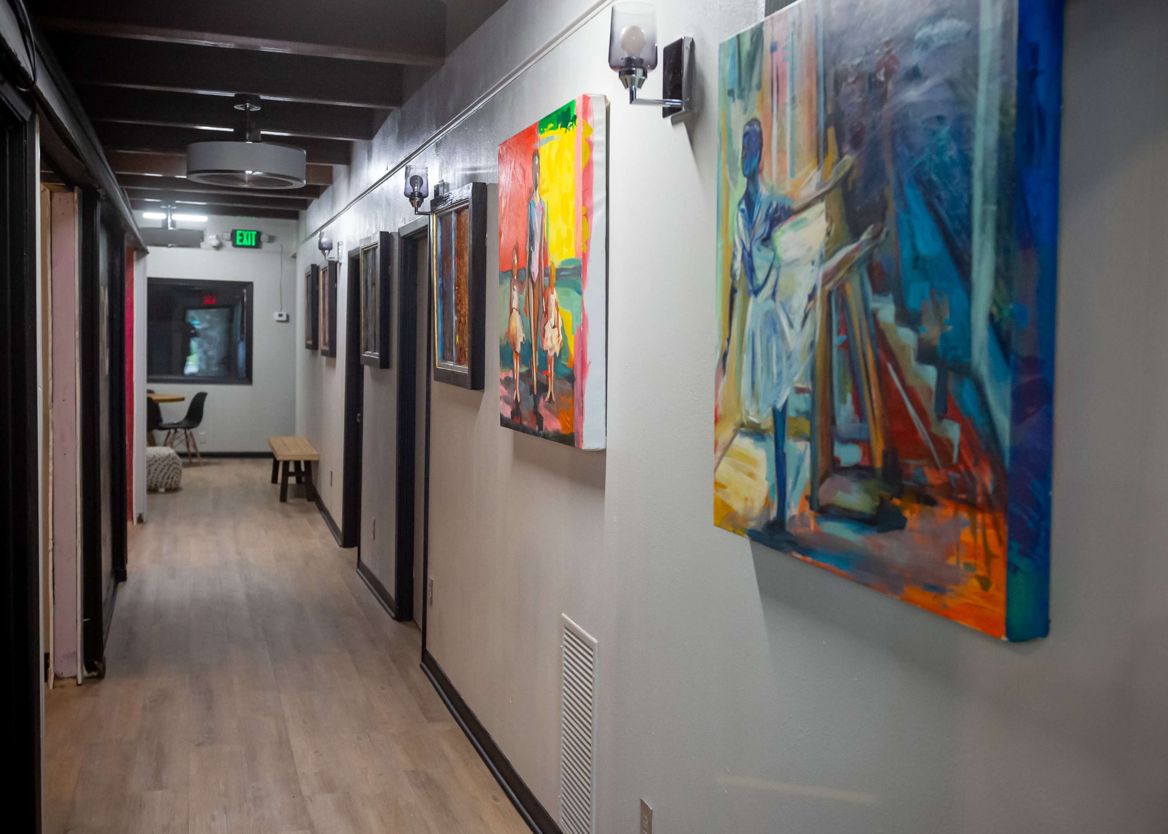 The image shows a well-lit corridor featuring several colorful paintings on the walls, with wooden flooring and exit signs.