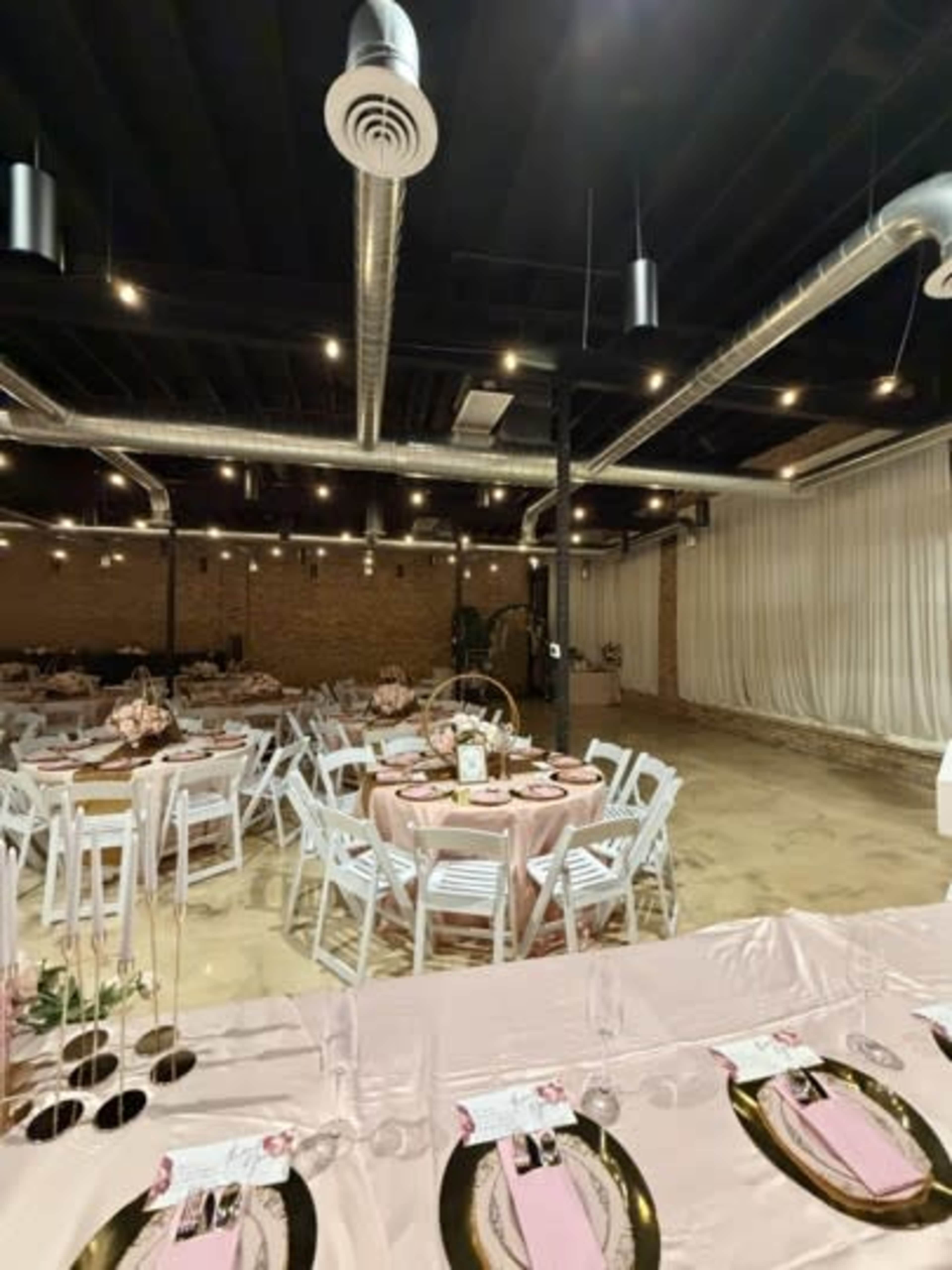 The image shows a modern event space with round tables covered in pink tablecloths, white chairs, and decorative centerpieces, featuring exposed brick walls and industrial lighting.