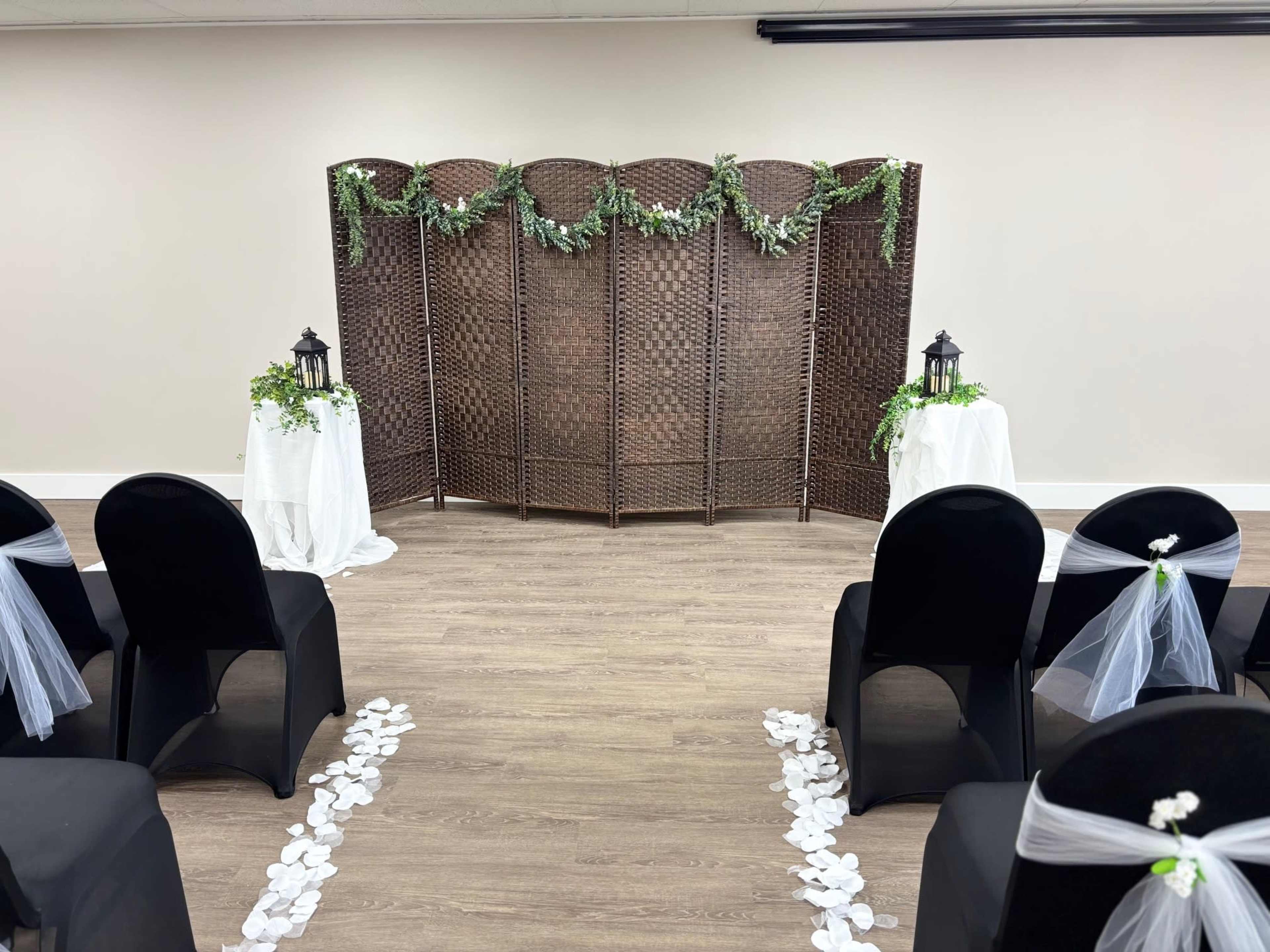 The image shows a decorated indoor space with a folding screen in the center, flanked by small tables with lanterns, surrounded by black chairs tied with white sashes and scattered flower petals on the floor.
