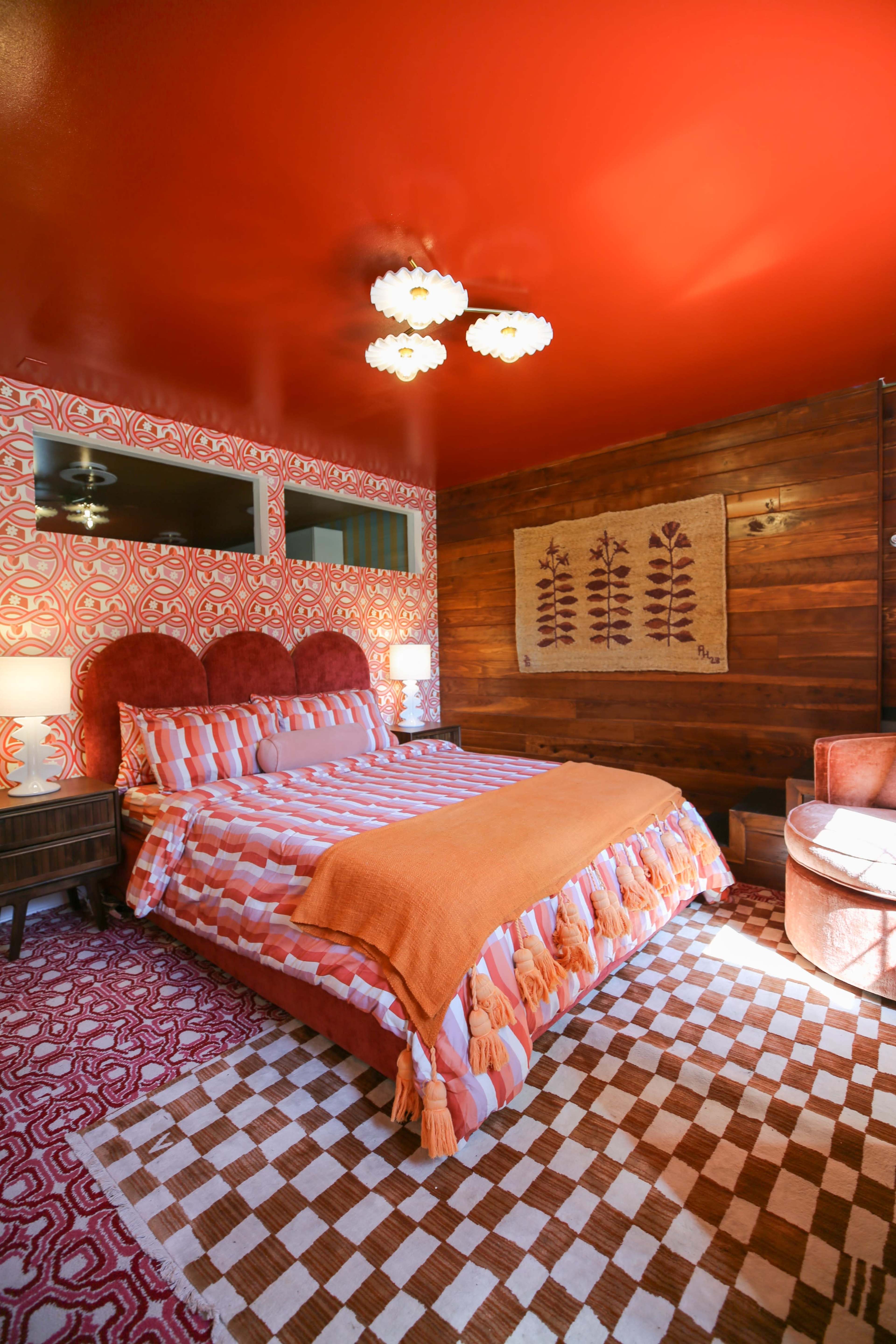 A brightly colored bedroom featuring a patterned bedspread, wooden accents, and a contemporary light fixture on a vivid orange wall.