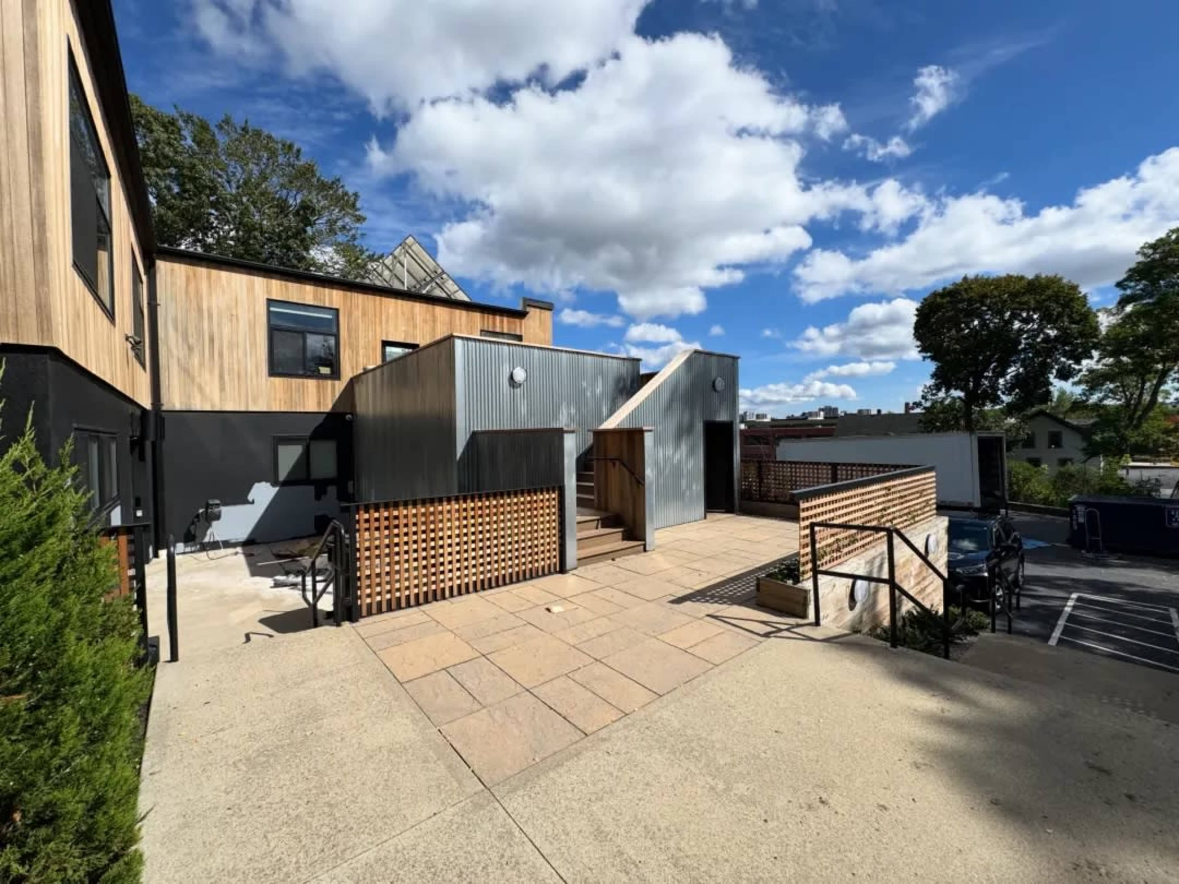 The image shows a modern building with a combination of wood and metal siding, featuring a staircase and outdoor seating area under a blue sky with scattered clouds.