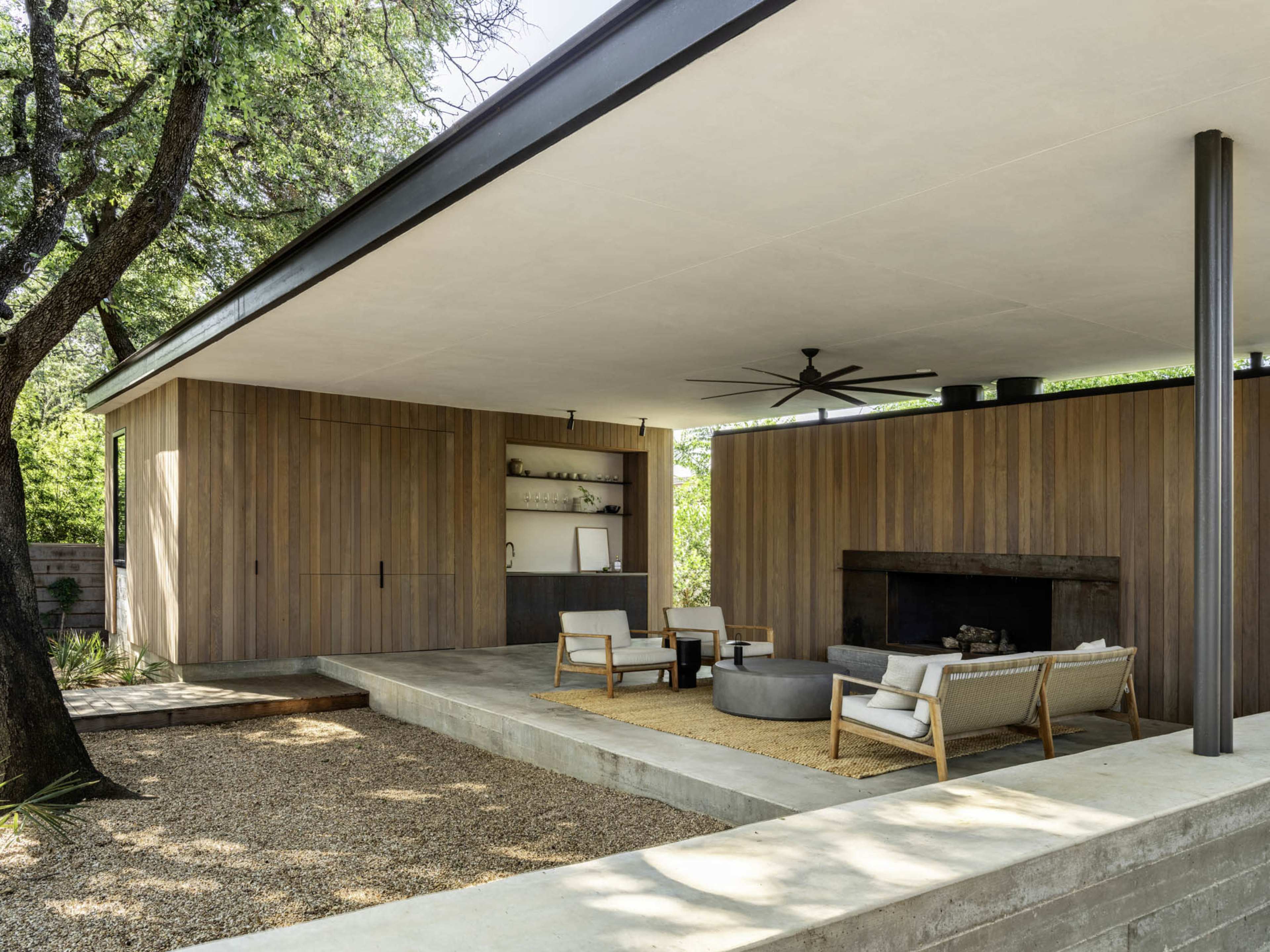 A modern outdoor living space features a wooden structure with a seating area, a fireplace, and a ceiling fan, surrounded by gravel and greenery.
