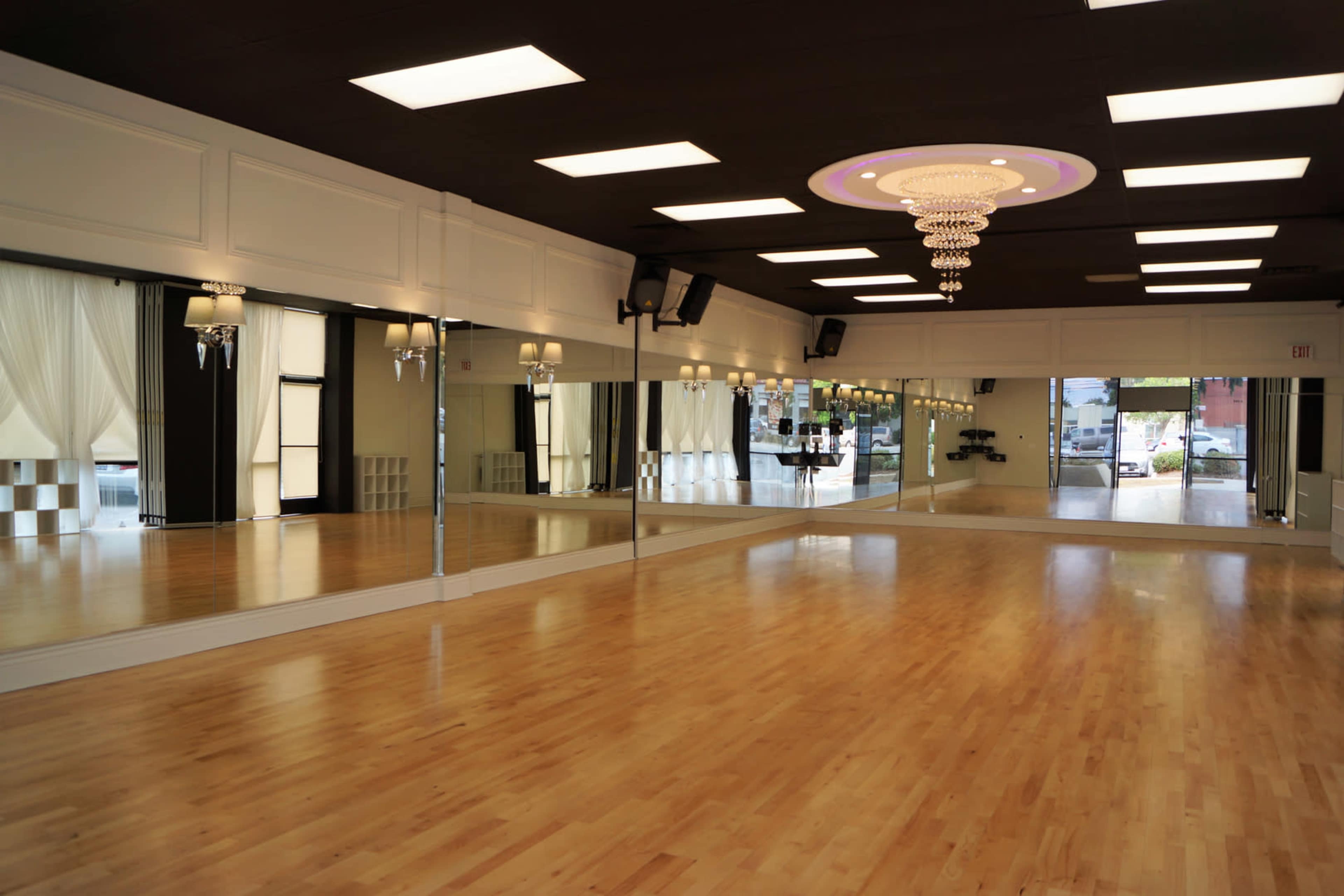 The image shows a spacious dance studio with hardwood floors, large mirrors on one wall, and a chandelier hanging from the ceiling.