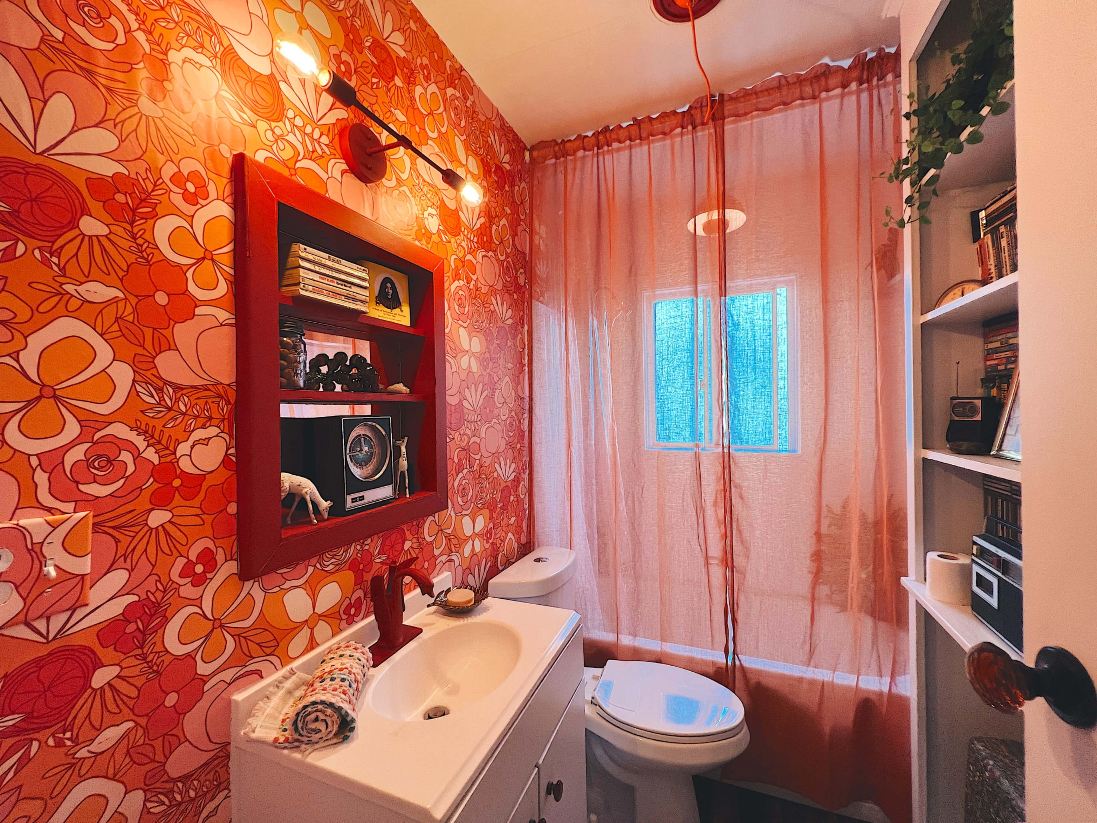 The image shows a small bathroom with floral-patterned wallpaper, a white vanity with a sink, a toilet, and a window covered by a sheer curtain.