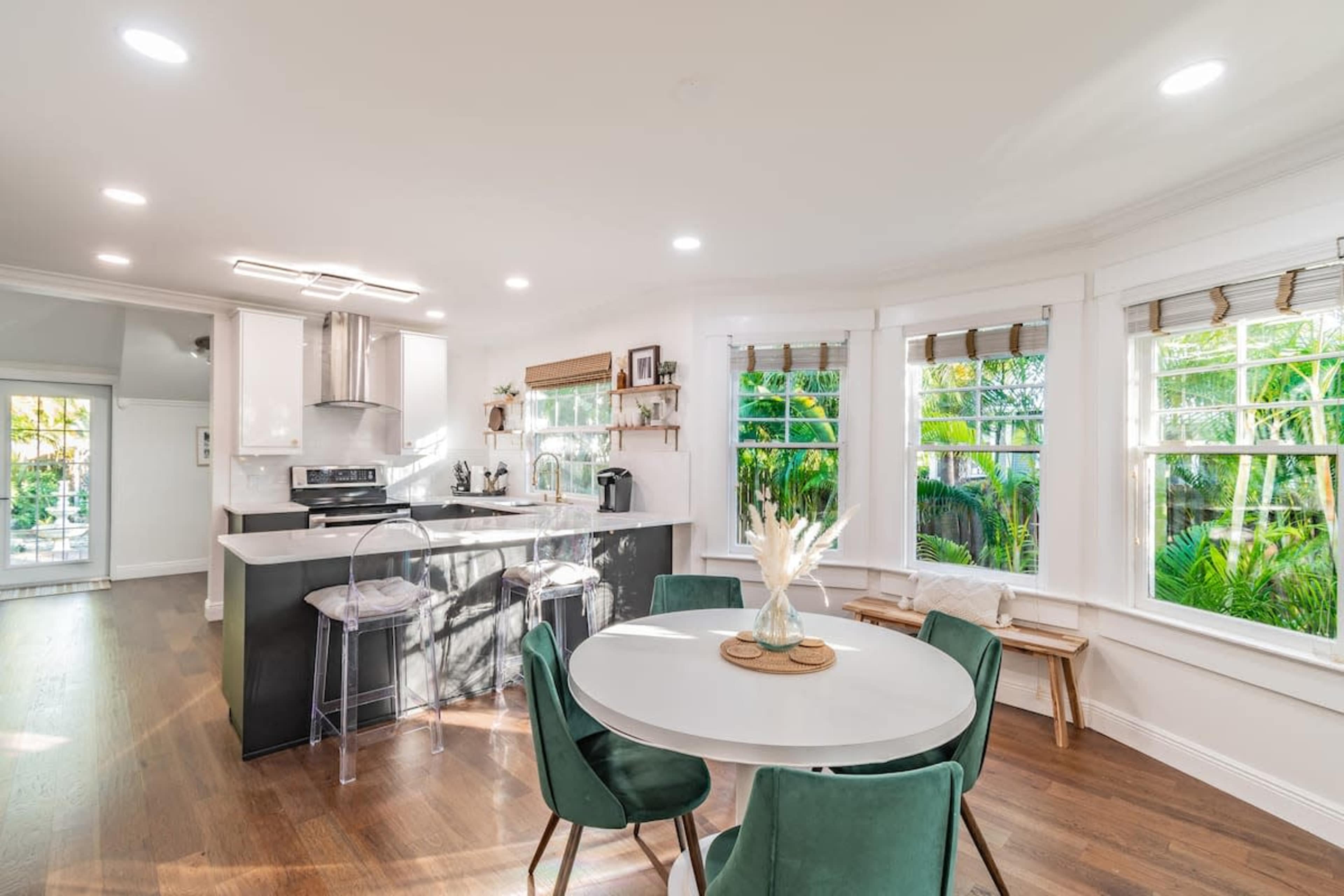 A bright, modern kitchen and dining area with a round table, green chairs, and large windows overlooking a garden.