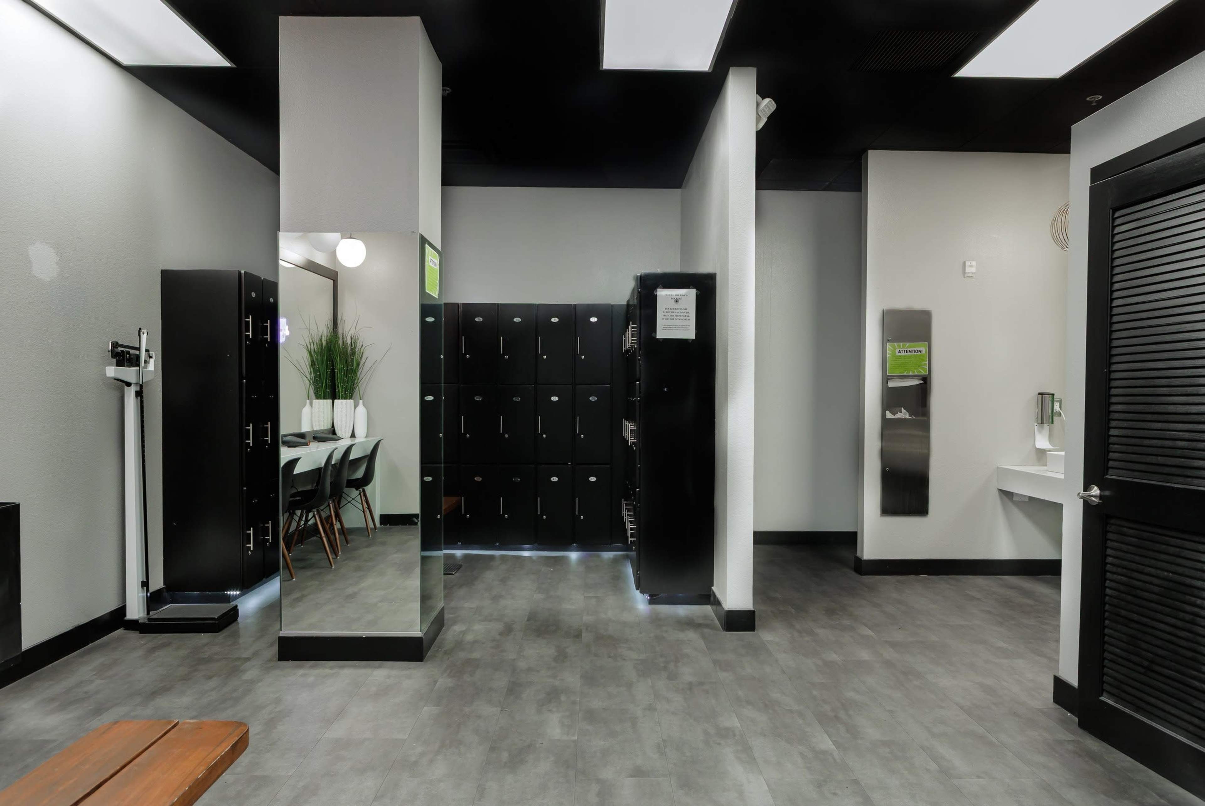 The image shows a modern changing room with black lockers, a mirrored section, and a seating area with a table and chairs.