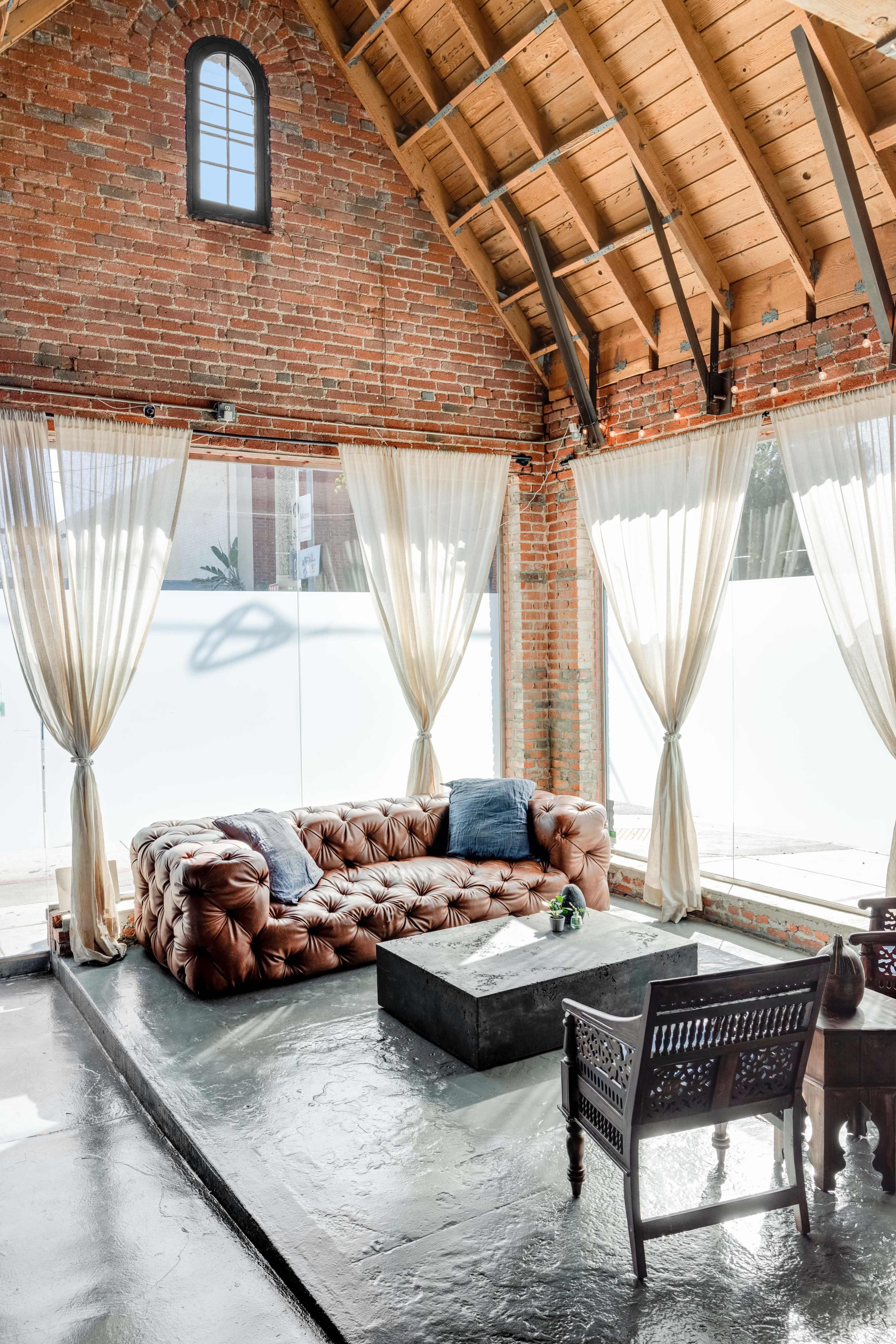 A spacious living area with a brown tufted leather sofa, a concrete coffee table, and large windows with sheer curtains.