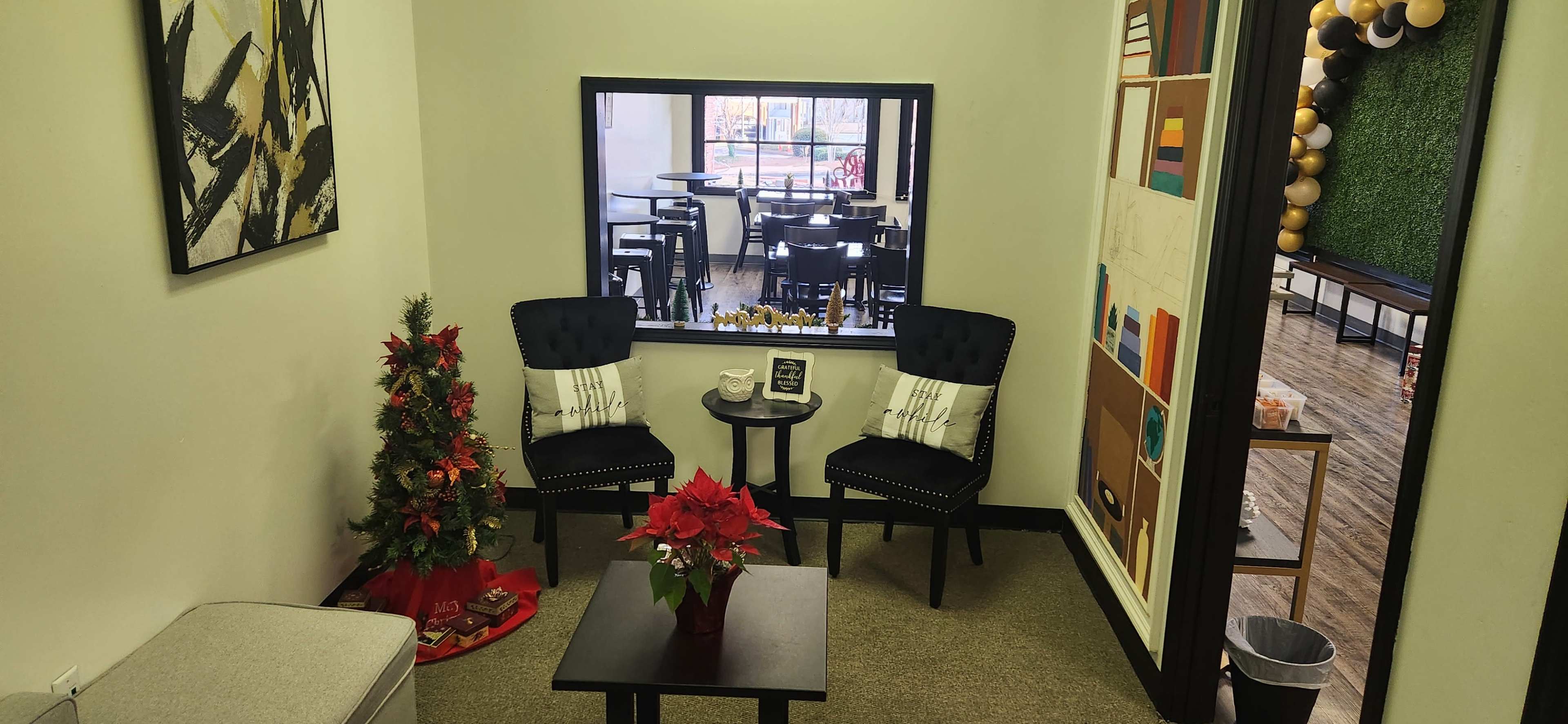 The image shows a small seating area with two black chairs and a table, decorated with a poinsettia and a small Christmas tree, next to a window overlooking a dining area.