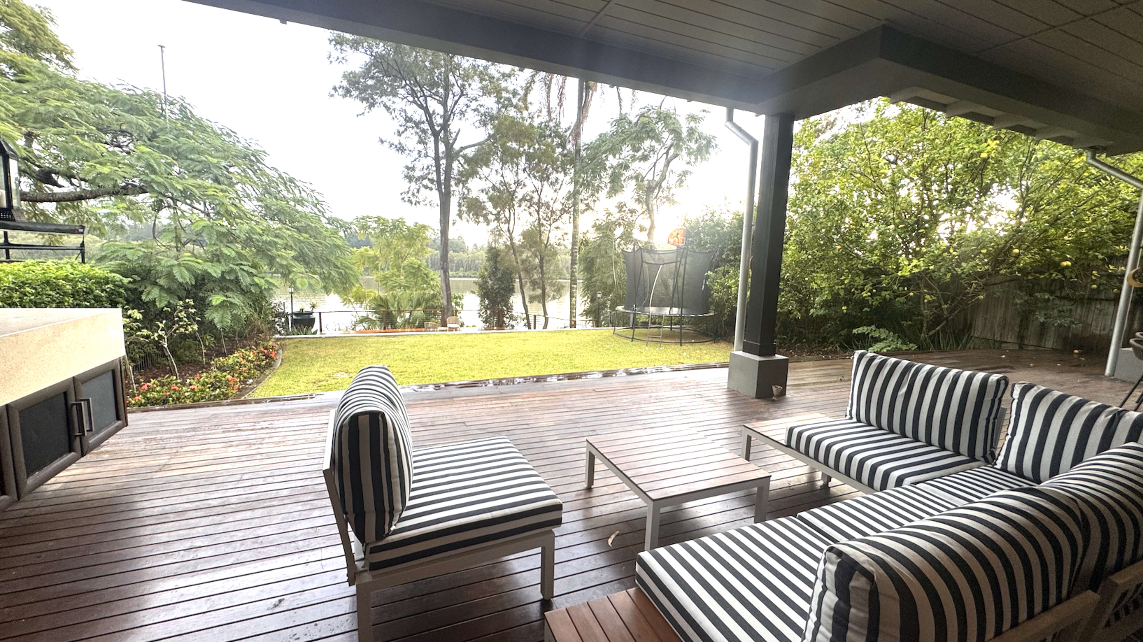 The image shows a spacious wooden deck with striped outdoor seating, adjacent to a green lawn and trees overlooking a body of water.