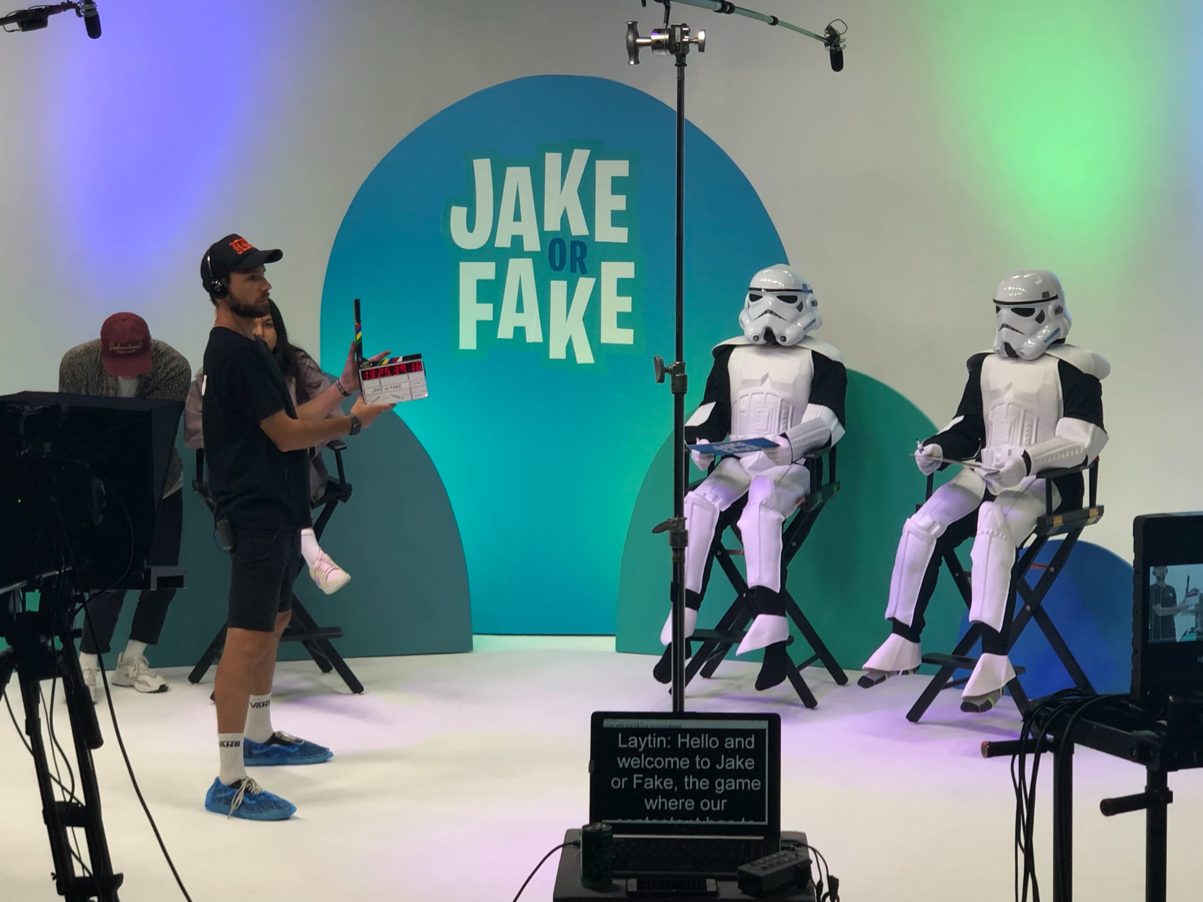 A person holds a clapperboard in front of two individuals dressed as stormtroopers, while a camera crew prepares for a production set against a colorful backdrop that reads "JAKE or FAKE."