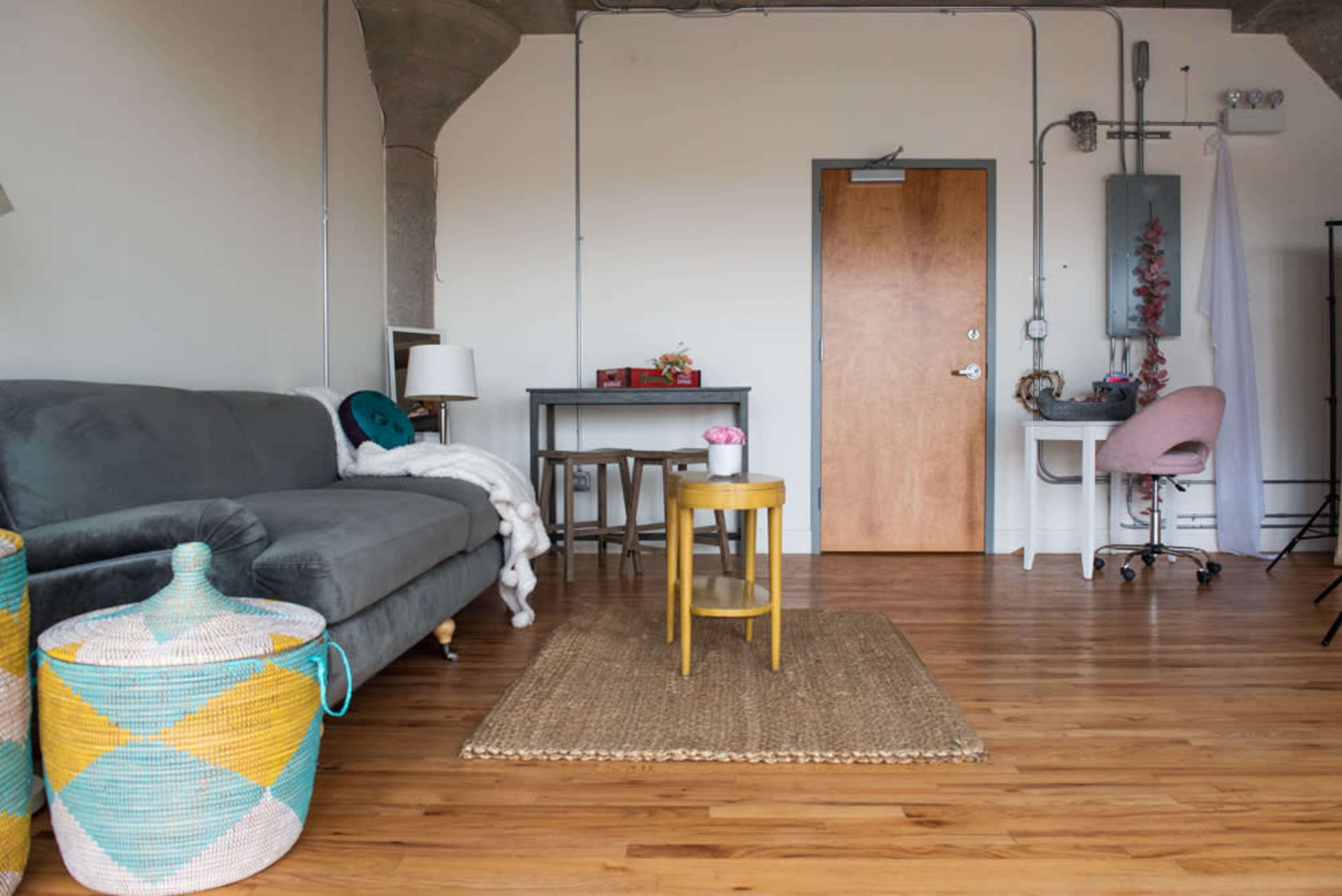 A minimalist living space featuring a gray sofa, a small round yellow table, and a desk with a pink chair, all arranged on a wooden floor.