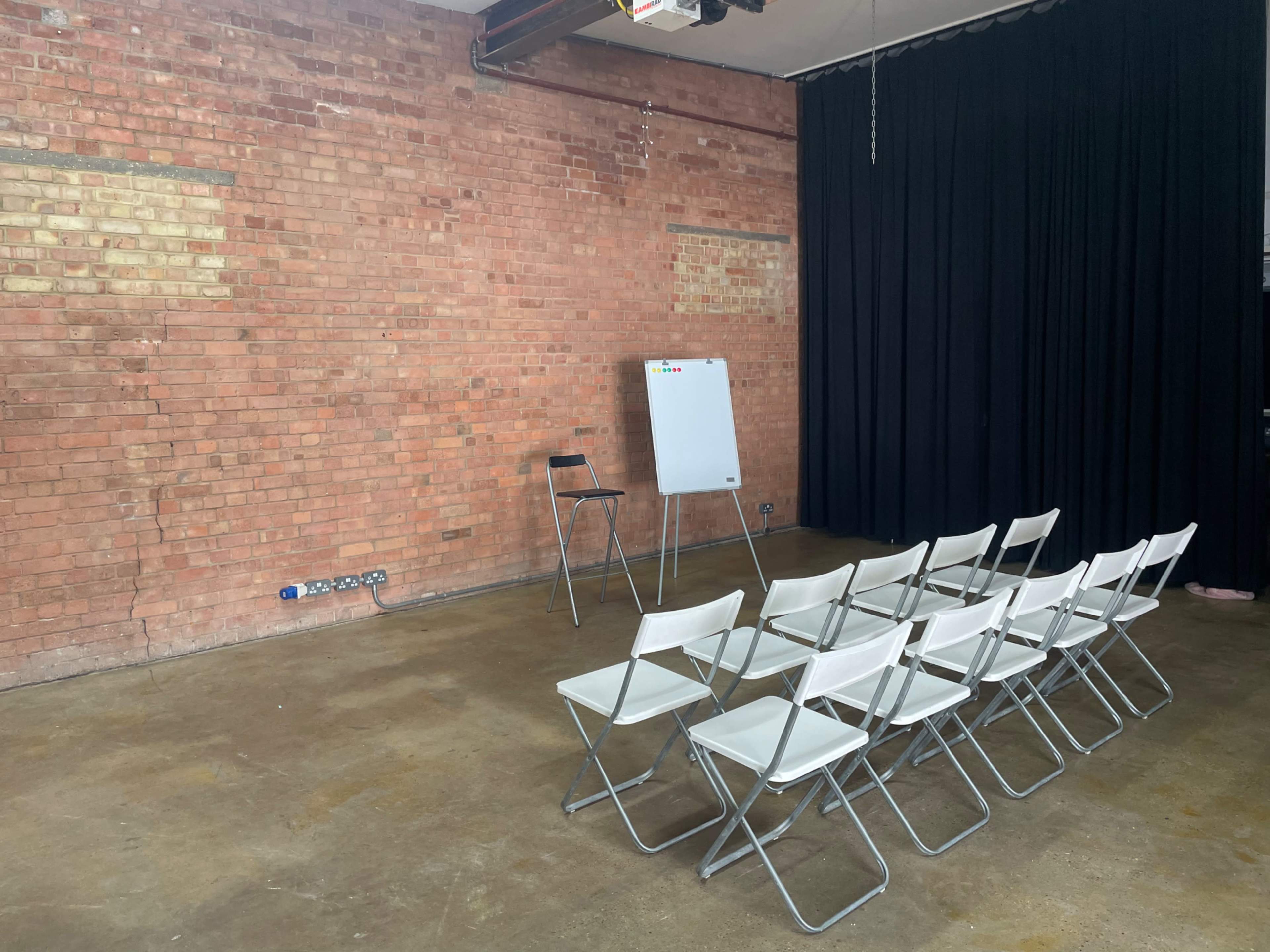A room with exposed brick walls features a row of white folding chairs facing a blank flip chart on a stand beside a single black chair.