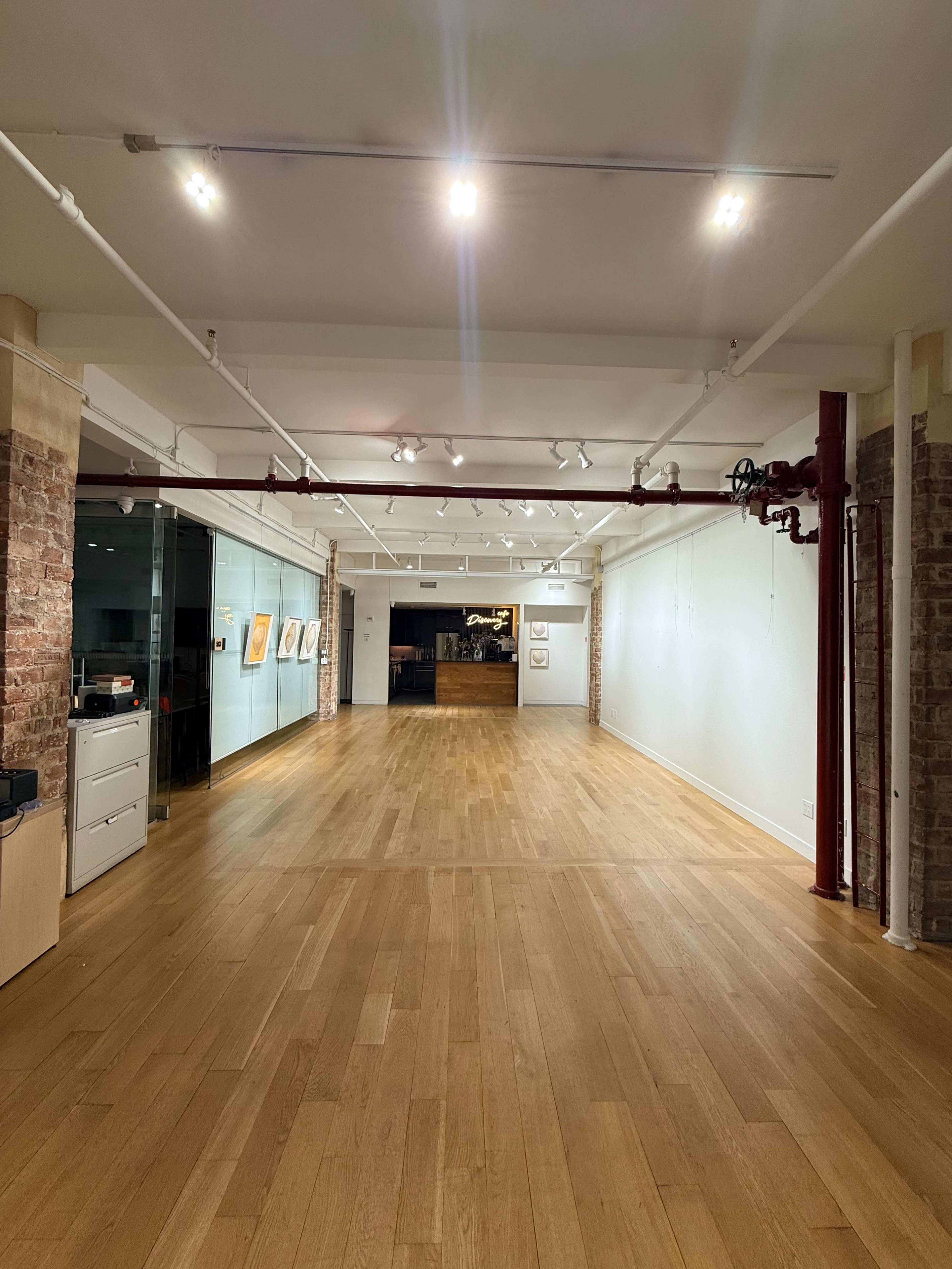 The image shows an empty gallery space with wooden flooring and exposed brick walls, featuring track lighting and an open layout leading to a darkened area in the background.
