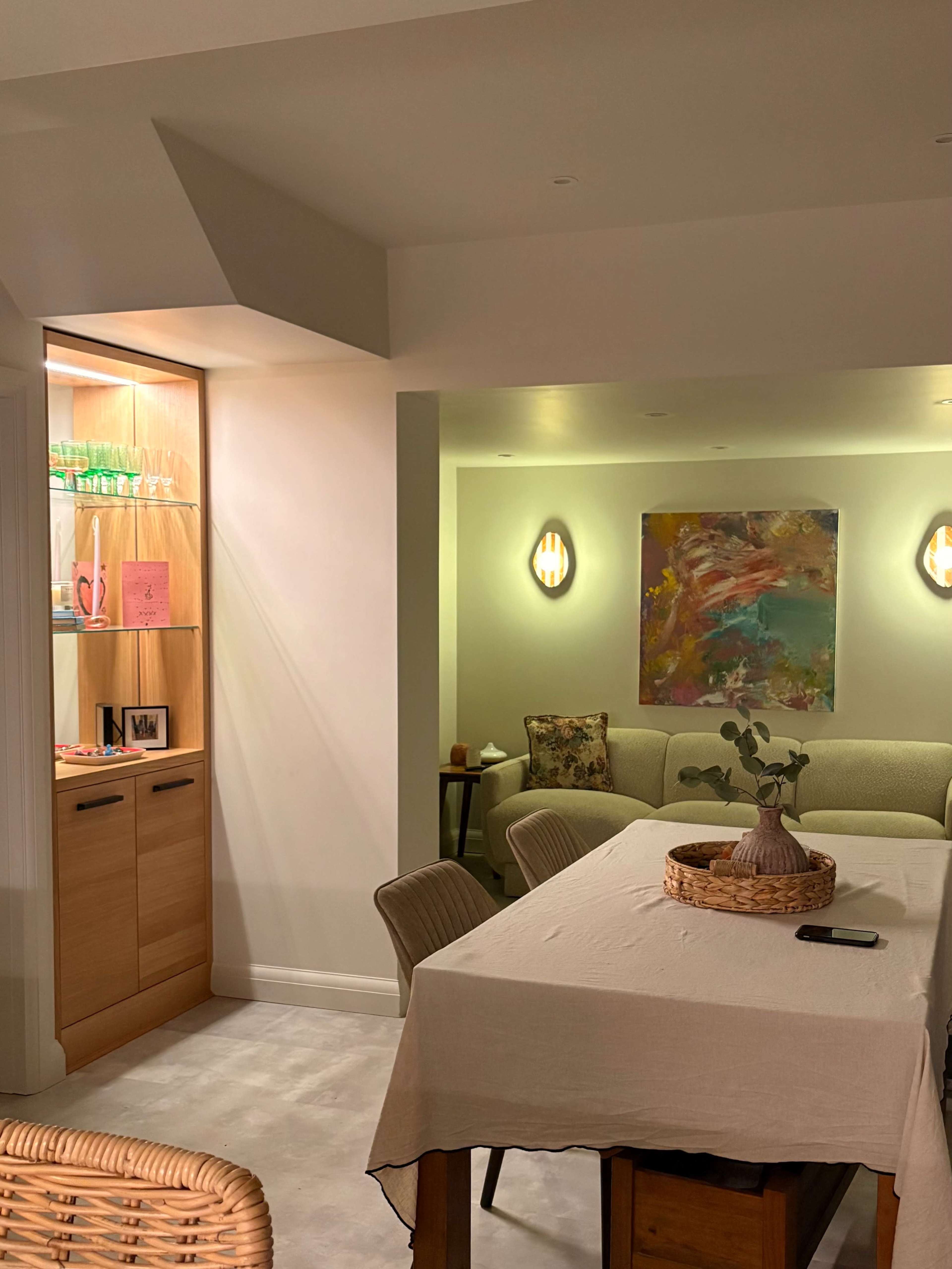 A well-lit dining area features a wooden table with a light-colored tablecloth, surrounded by chairs, and a display cabinet with glass shelves on one side.