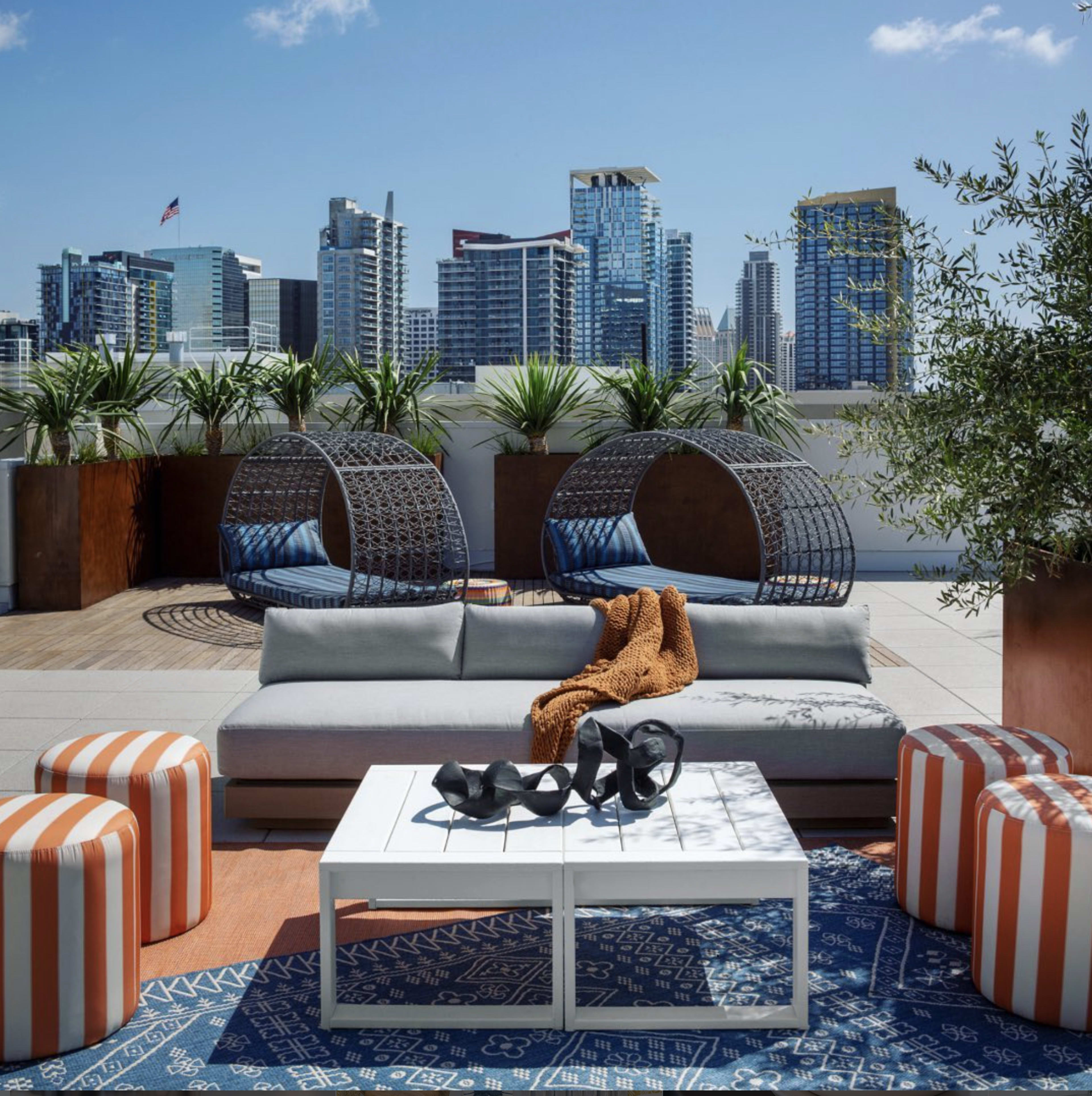 The image shows a modern rooftop lounge area with comfortable seating, decorative plants, and a city skyline in the background.