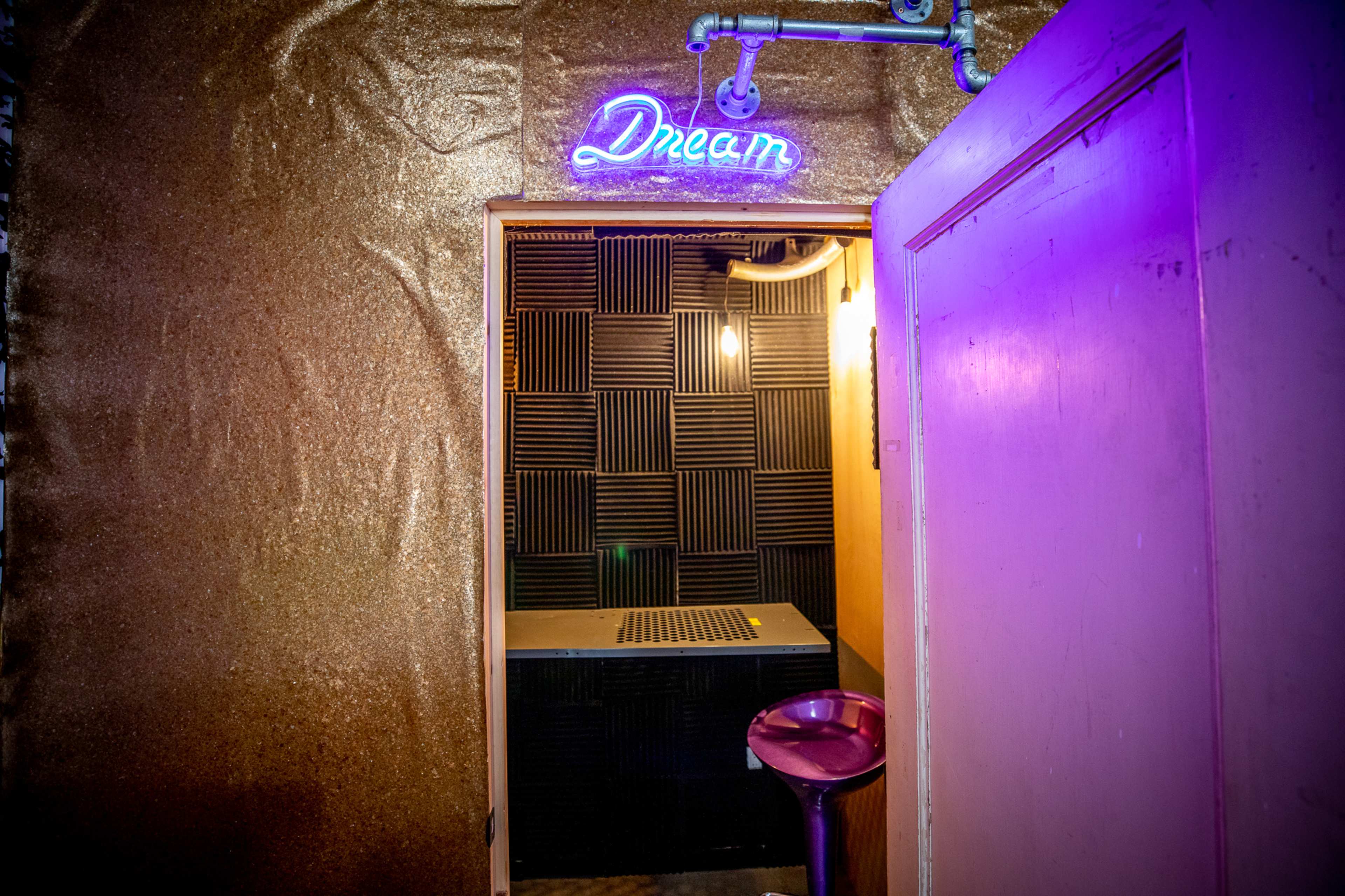 The image shows an entrance to a small room with a "Dream" neon sign, a dark textured wall, and a single chair beside a small table.