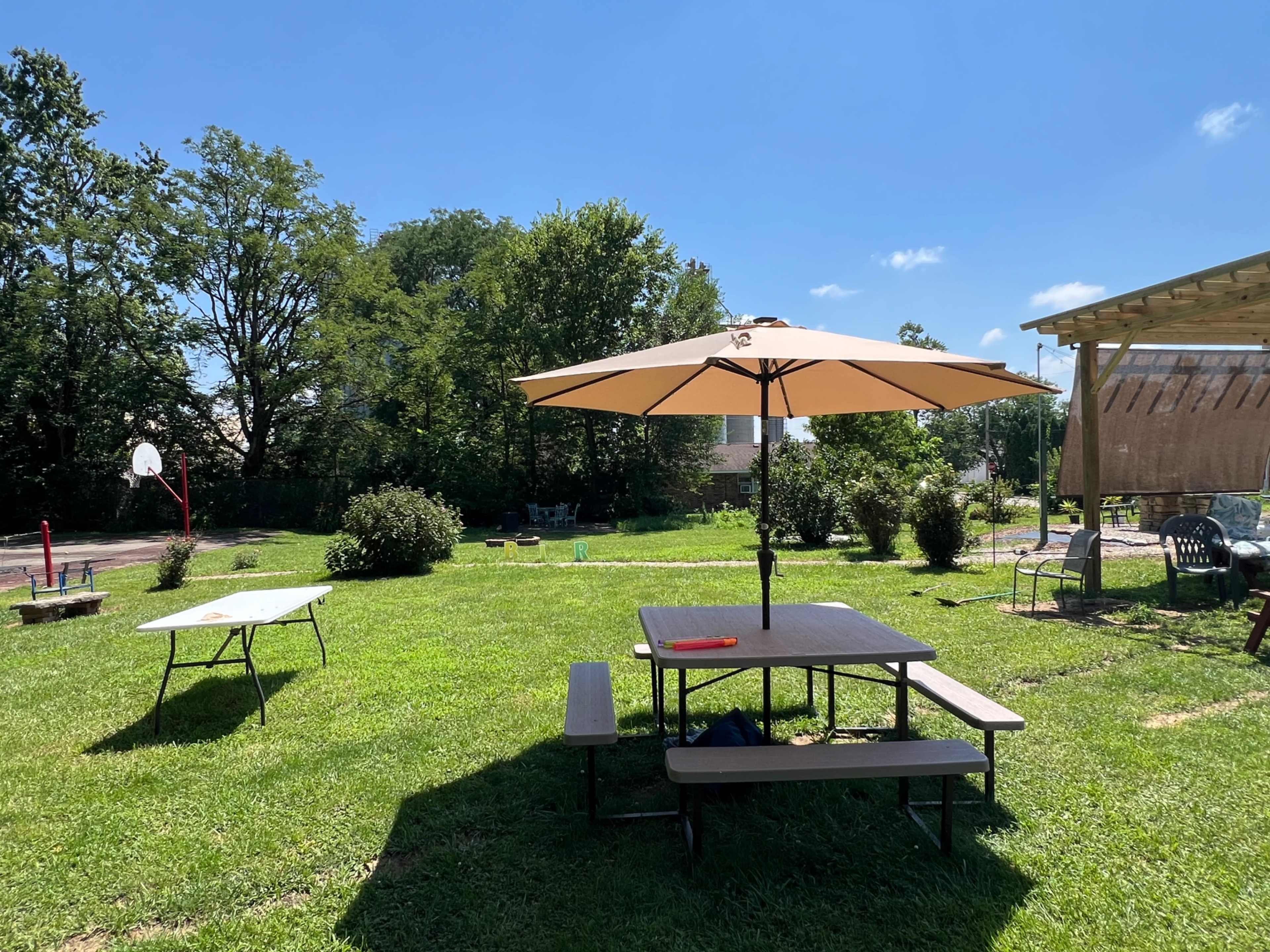 The image shows a grassy backyard with a picnic table under an umbrella, a basketball hoop in the background, and a few chairs scattered around the area.