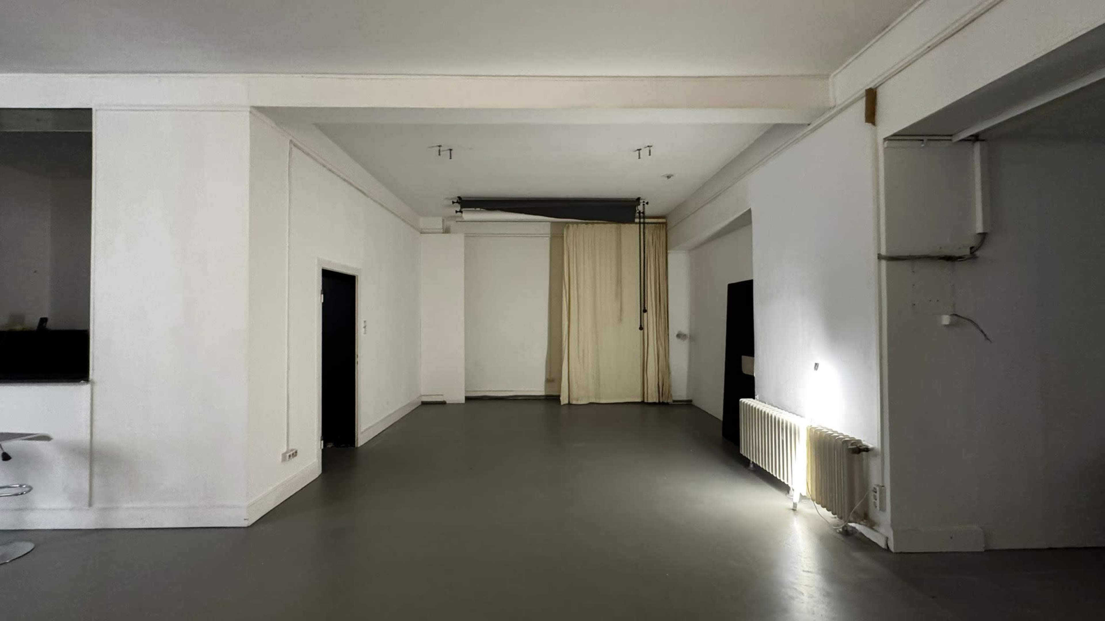 The image shows a spacious, empty room with white walls, a concrete floor, and a curtain partially drawn across an opening.