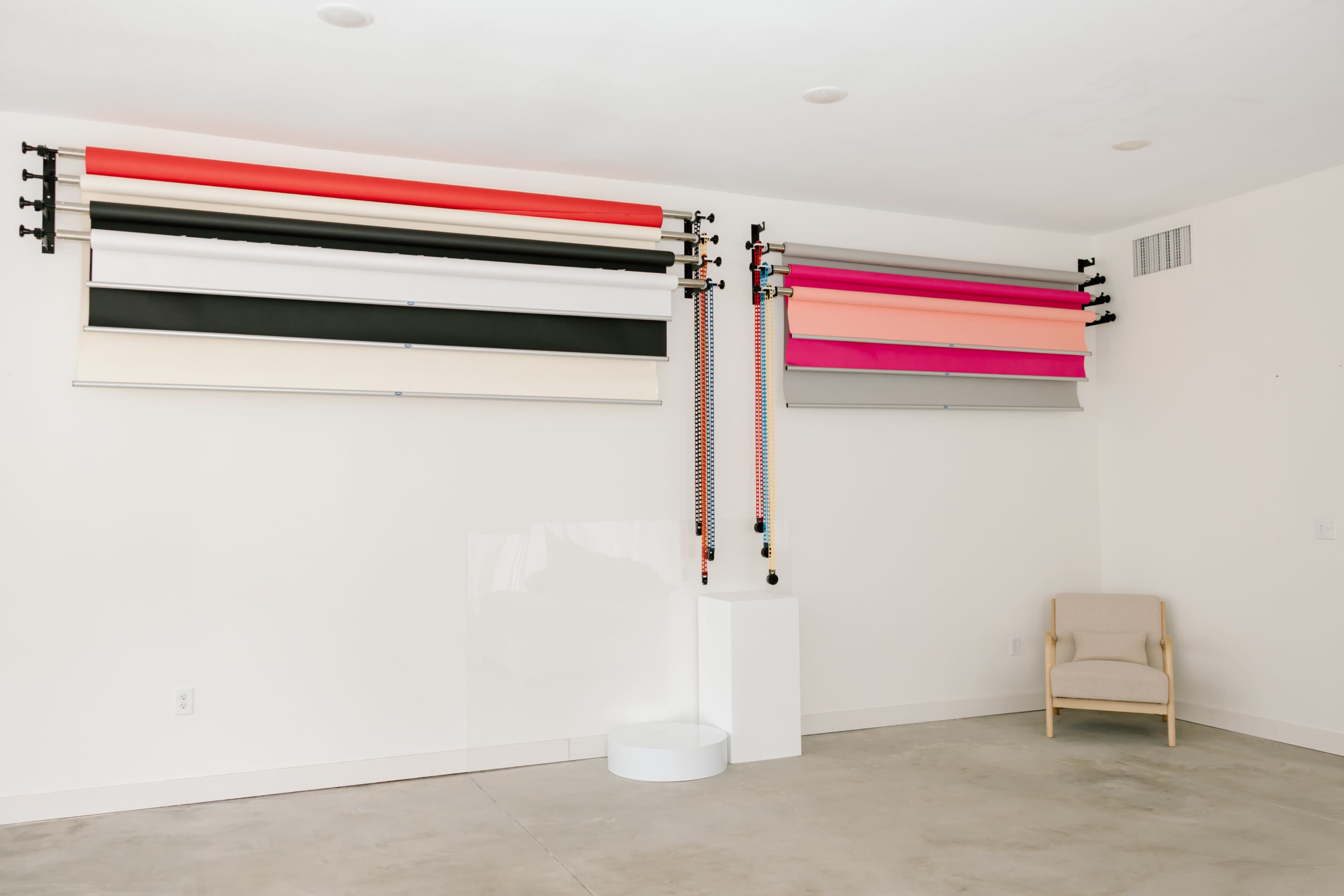 The image shows a minimalist room with multiple color backdrop rolls mounted on the wall and a single beige chair in the corner.