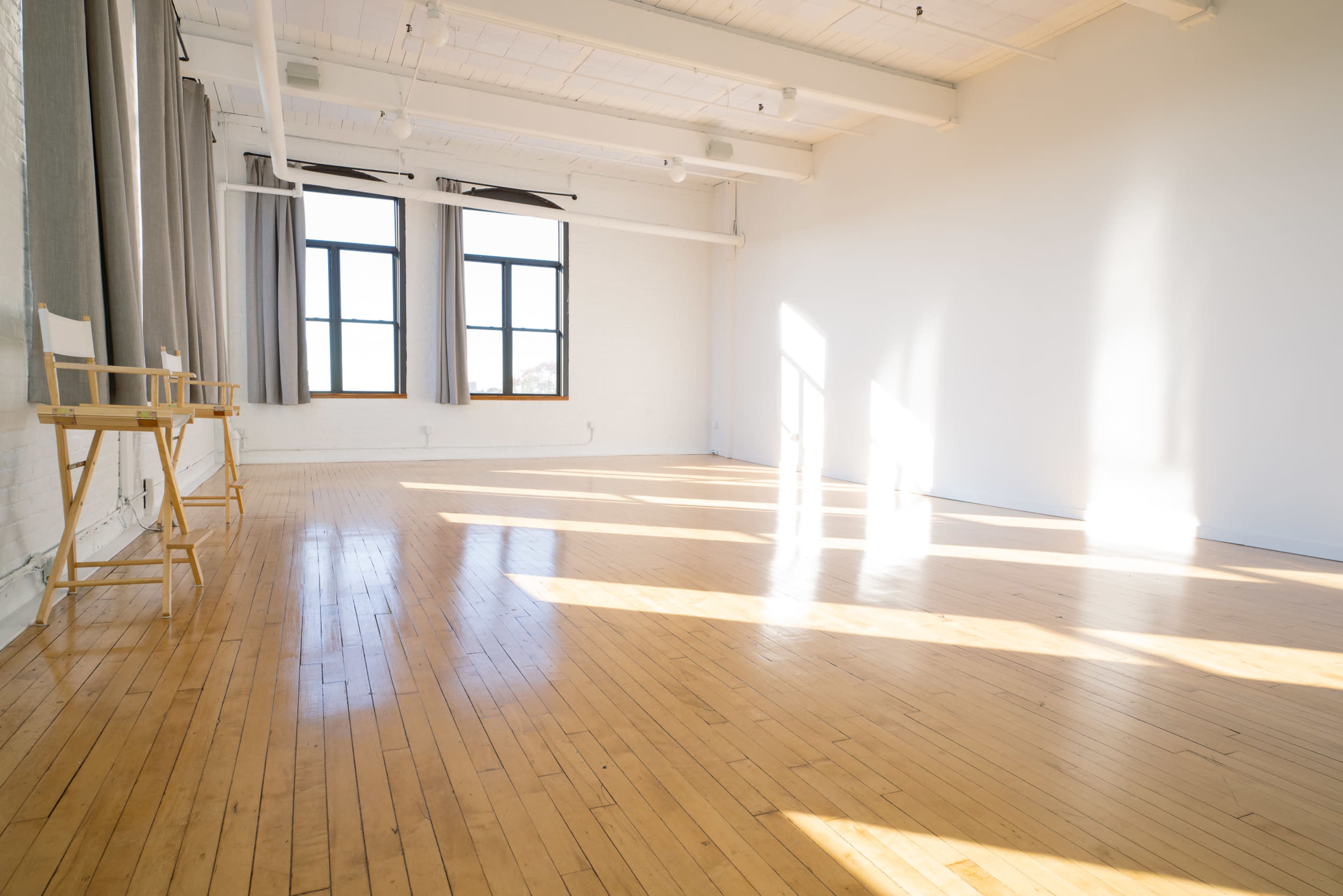 The image shows a spacious, empty room with wooden floors, large windows allowing natural light to stream in, and minimal furnishings.