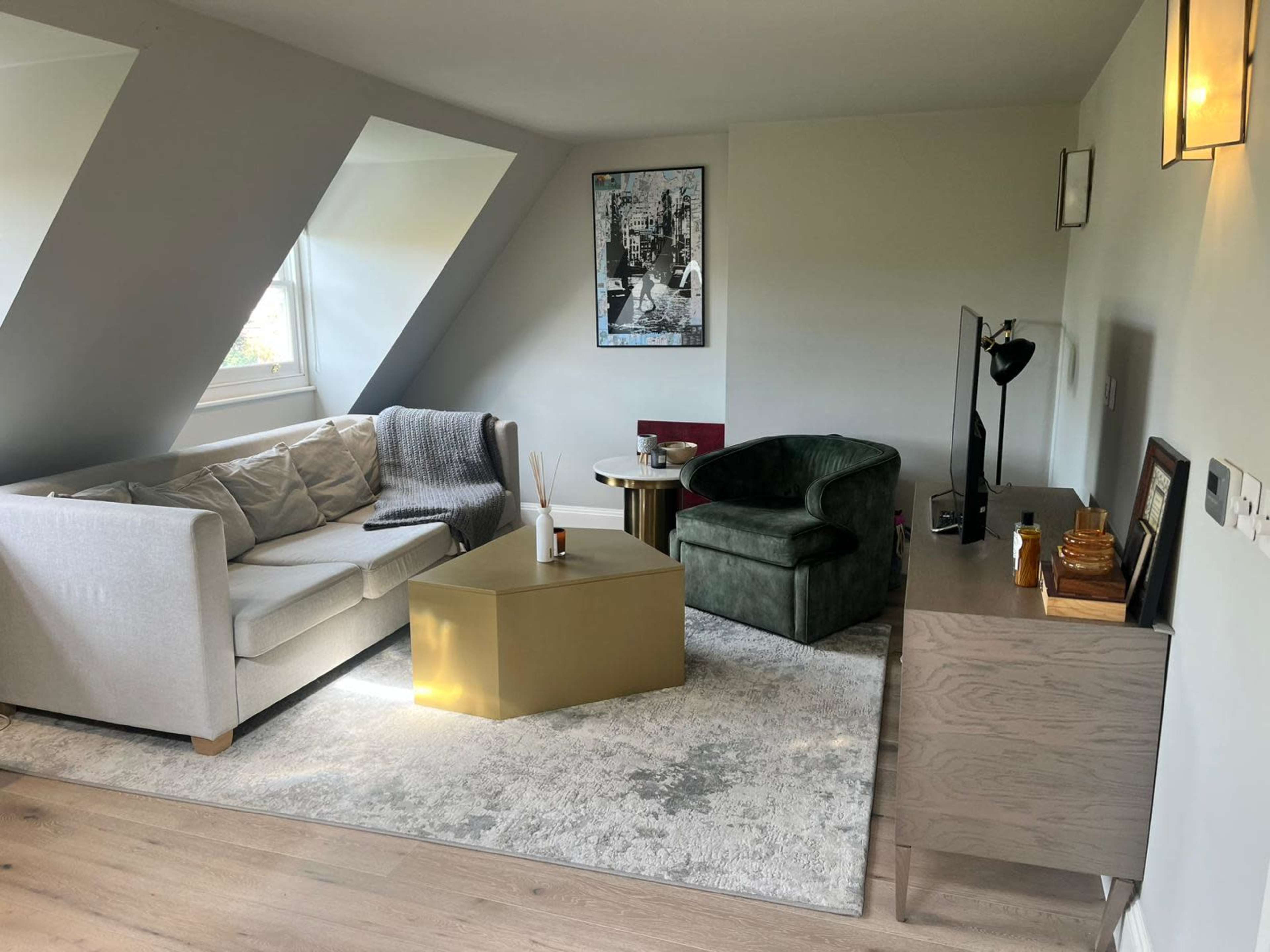 The image shows a modern living room with a light gray sofa, a green armchair, a round coffee table, and a wooden dresser, all arranged on a light rug under slanted walls.