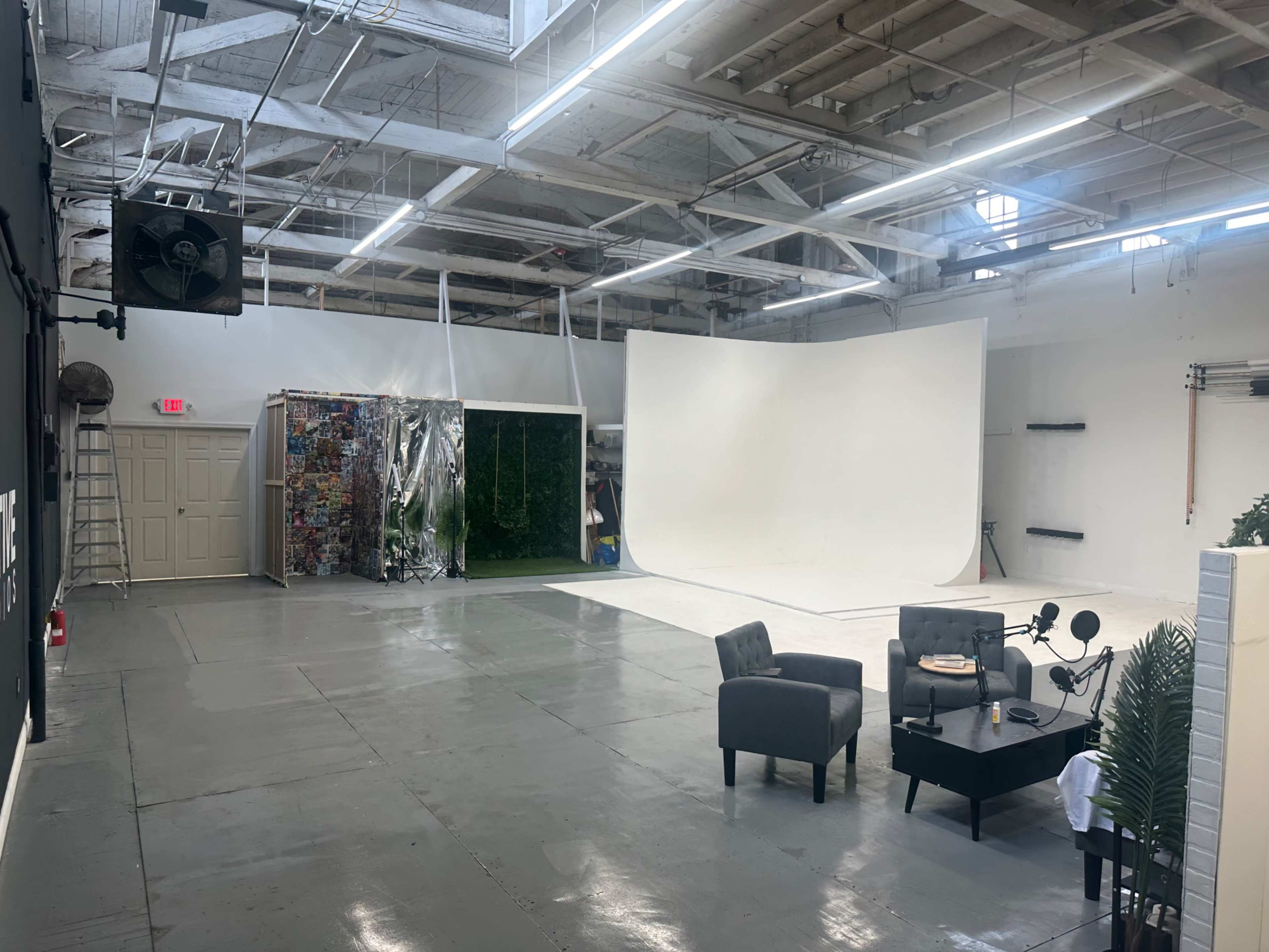 The image shows an empty photography studio with a white backdrop, various props, and two gray chairs arranged around a small table.