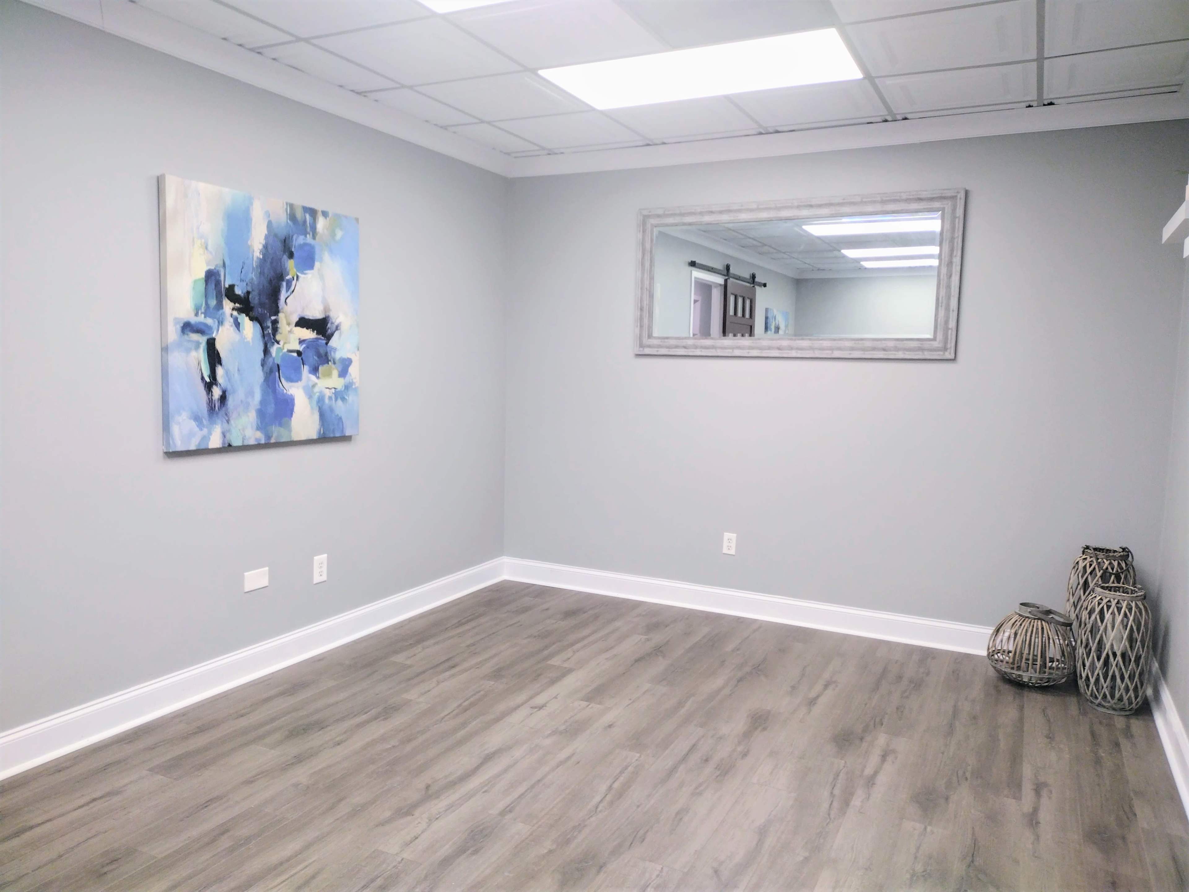 A light gray room features a large abstract painting on one wall, a rectangular mirror on the opposite wall, and two decorative baskets in the corner.