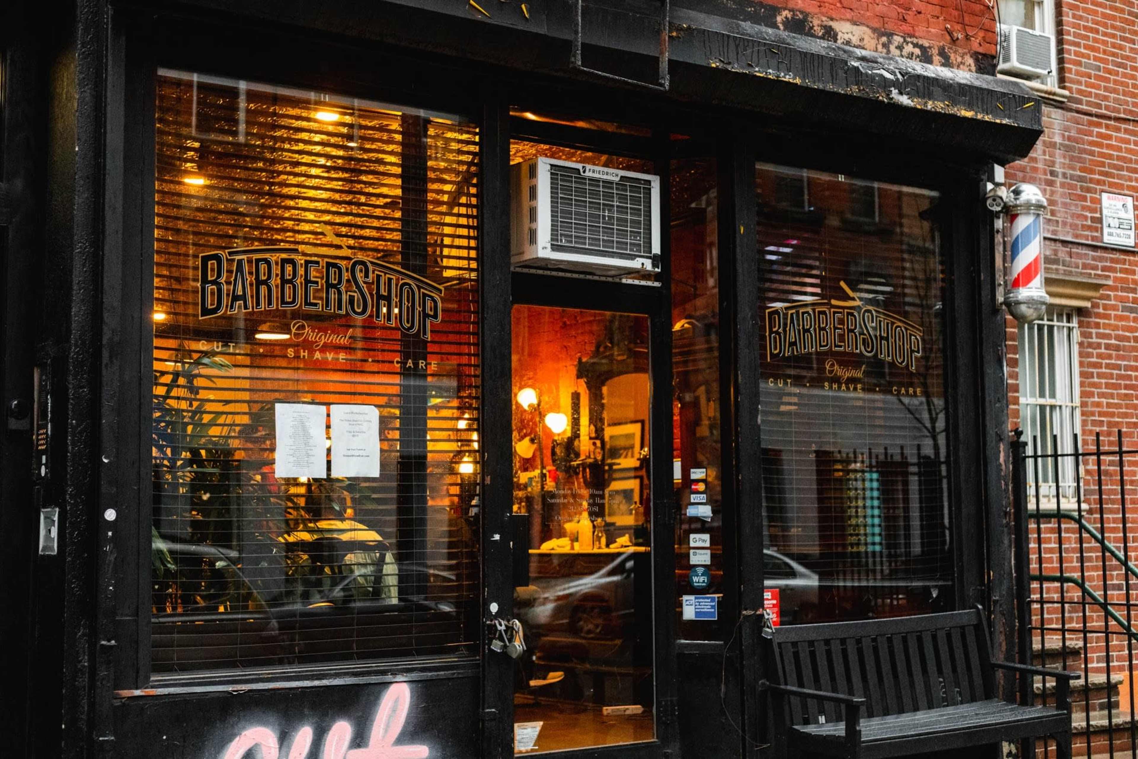 The storefront of a barbershop features large glass windows with signage, displaying the establishment's name and a view of the interior lit up warmly.