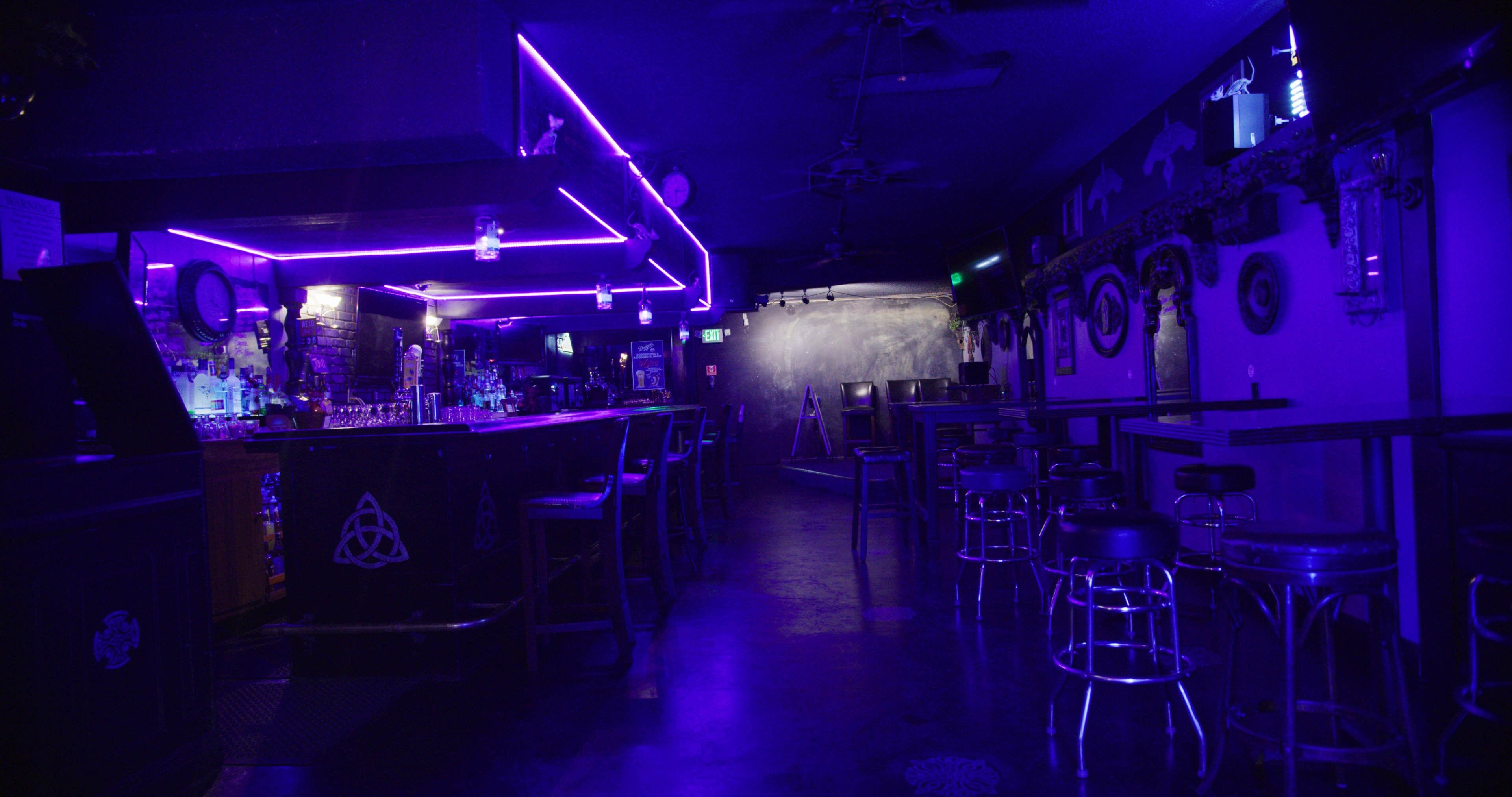 A dimly lit bar features a counter with glowing purple neon lights and several empty stools.