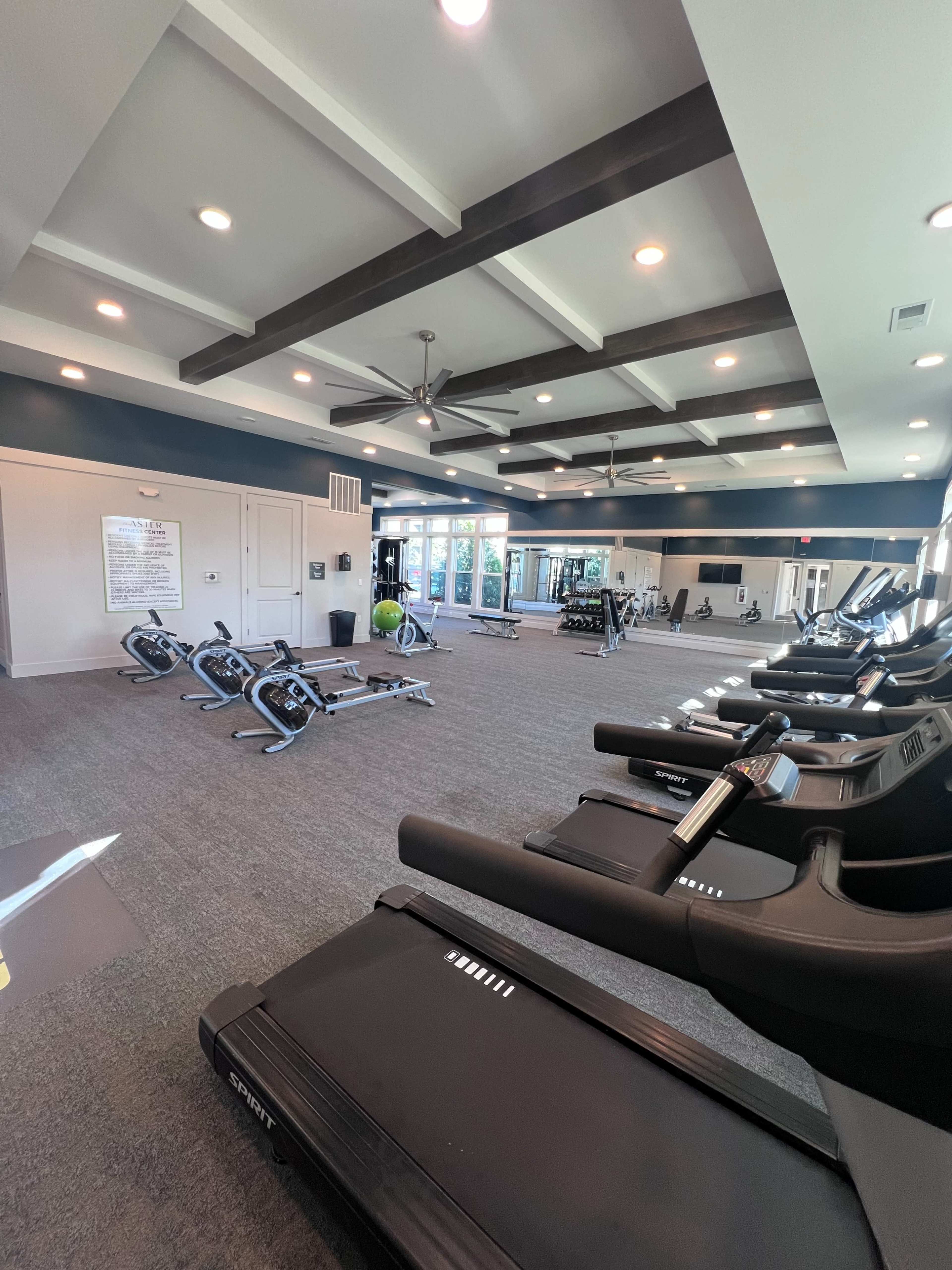 The image shows a spacious gym area equipped with treadmills, exercise bikes, and weightlifting equipment, illuminated by overhead lights.