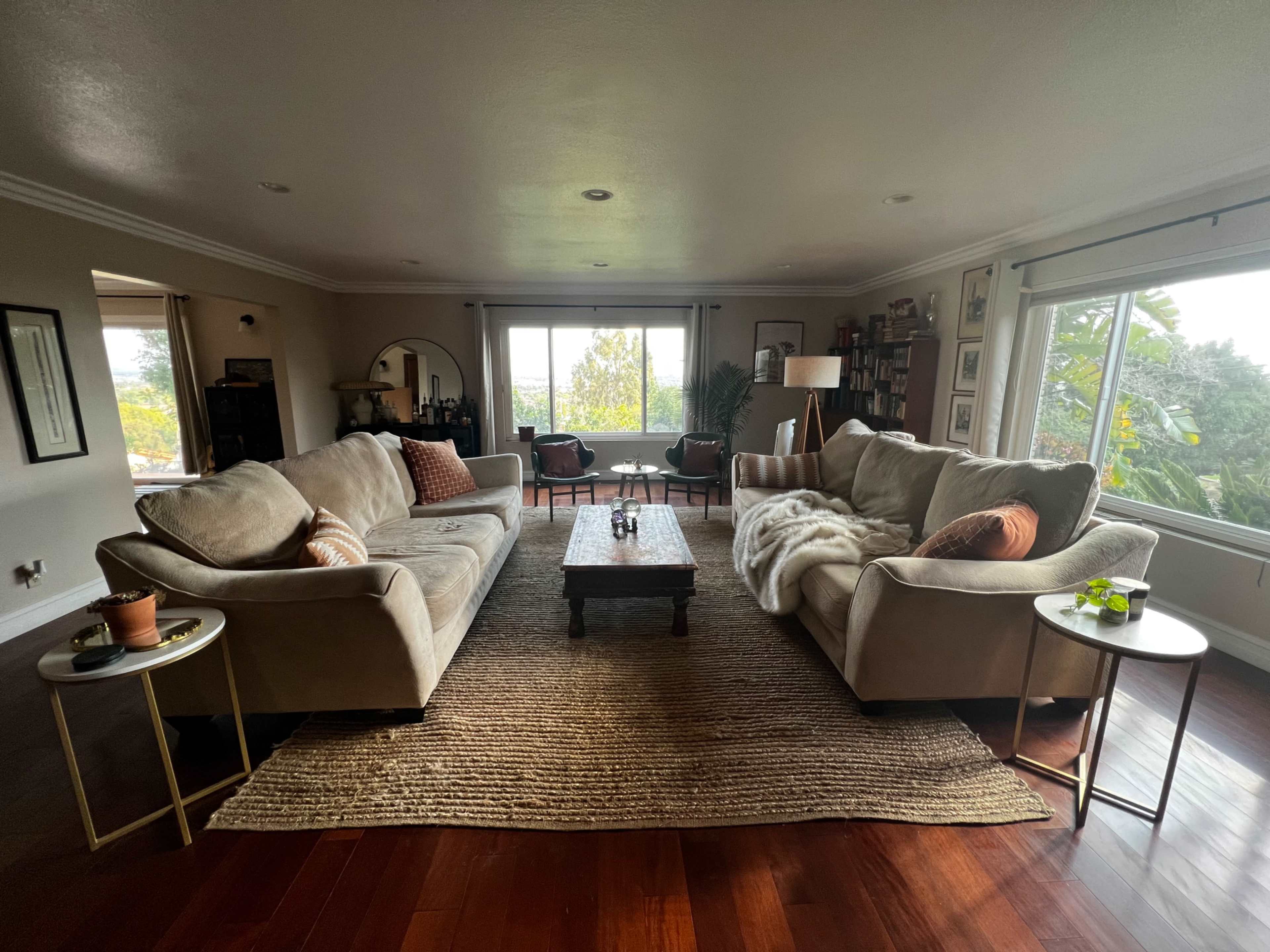 A spacious living room features two beige sofas facing a wooden coffee table, with large windows showcasing outdoor greenery.