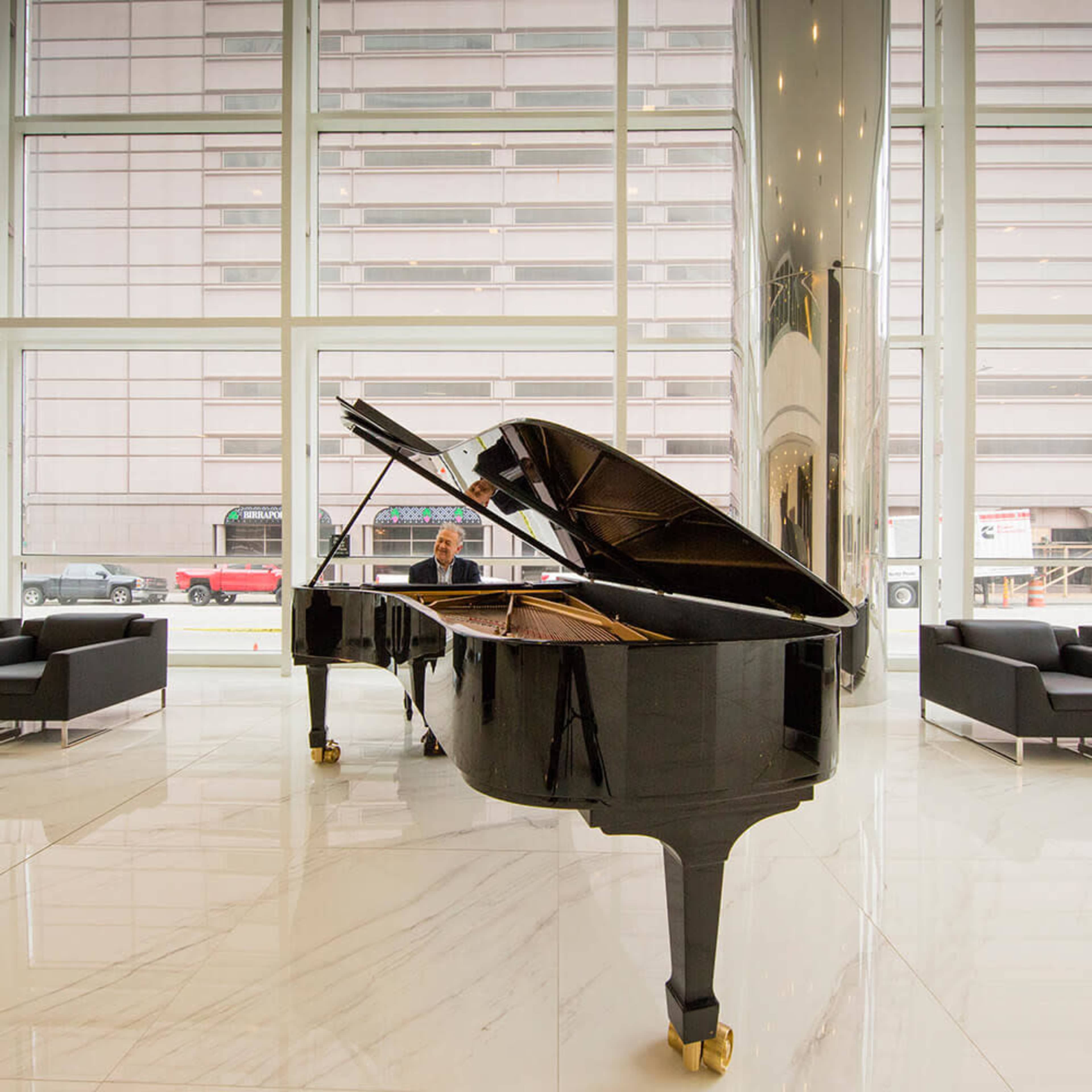 A grand piano is positioned in a modern, brightly lit lobby, with a person seated at the instrument and large windows displaying a cityscape in the background.