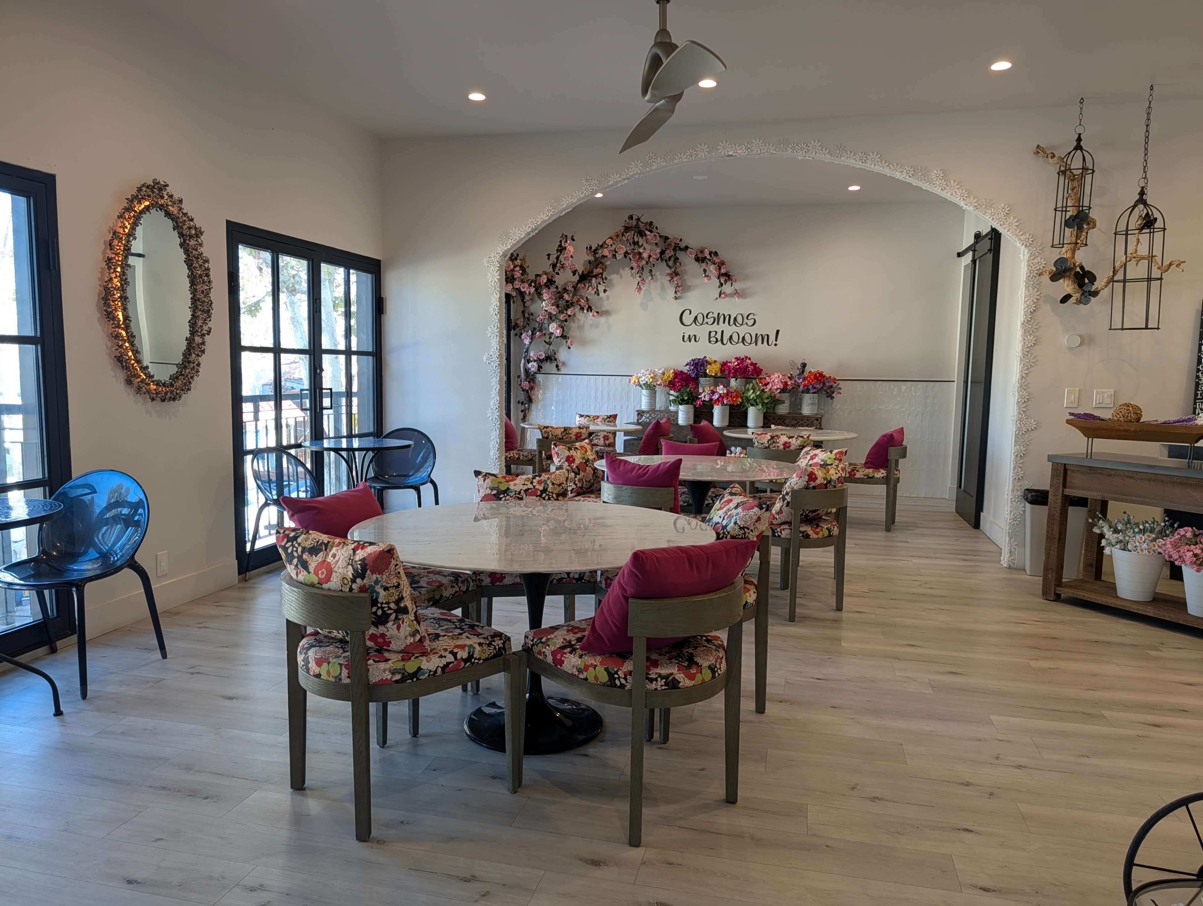 The image shows a bright, modern dining area with floral-patterned chairs, a large round table, and decorative elements including a wall with the phrase "Cosmos in Bloom" and potted flowers.