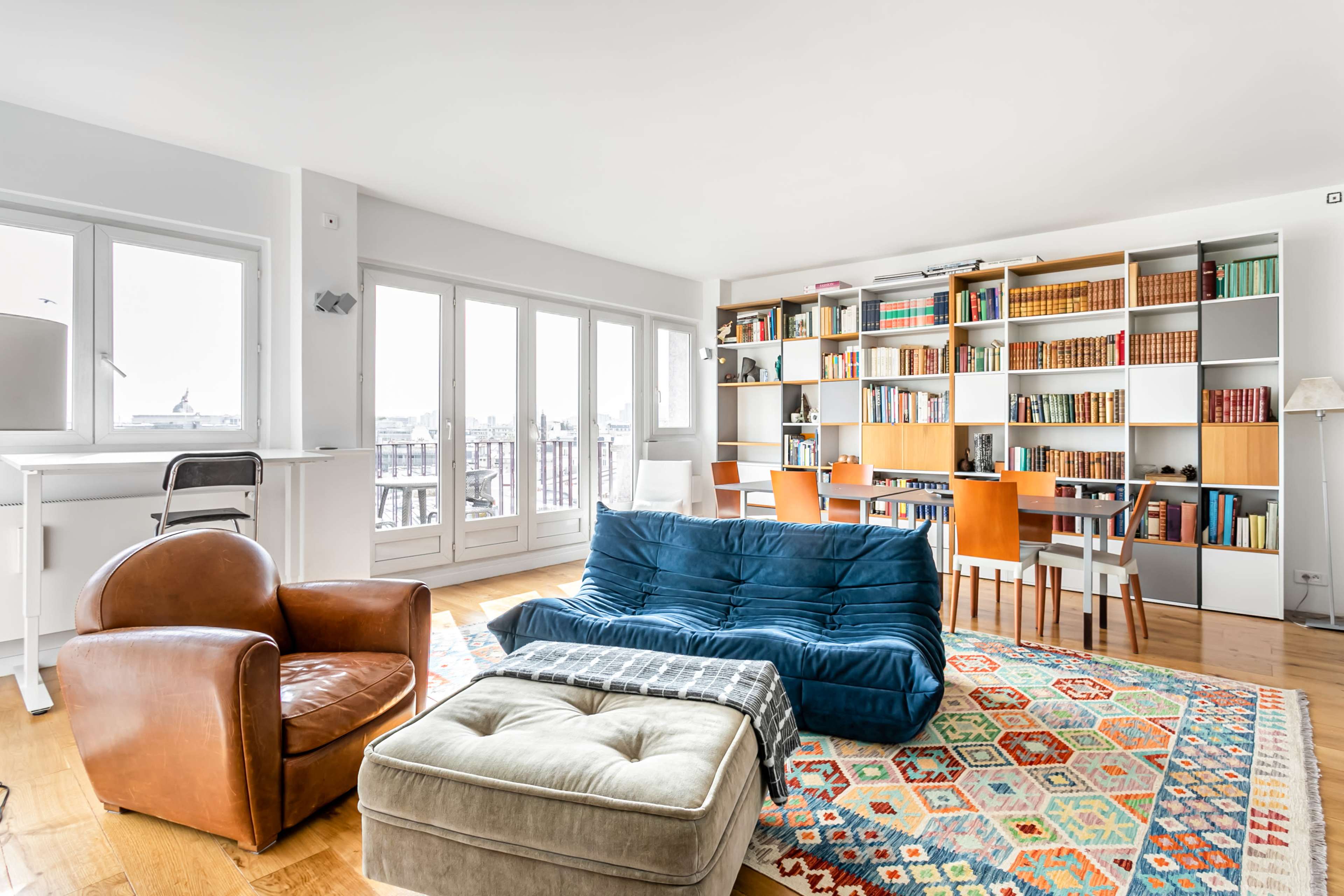 The image shows a modern living room with a blue sofa, an armchair, a large ottoman, and a bookshelf, along with a workspace and dining area by the windows.