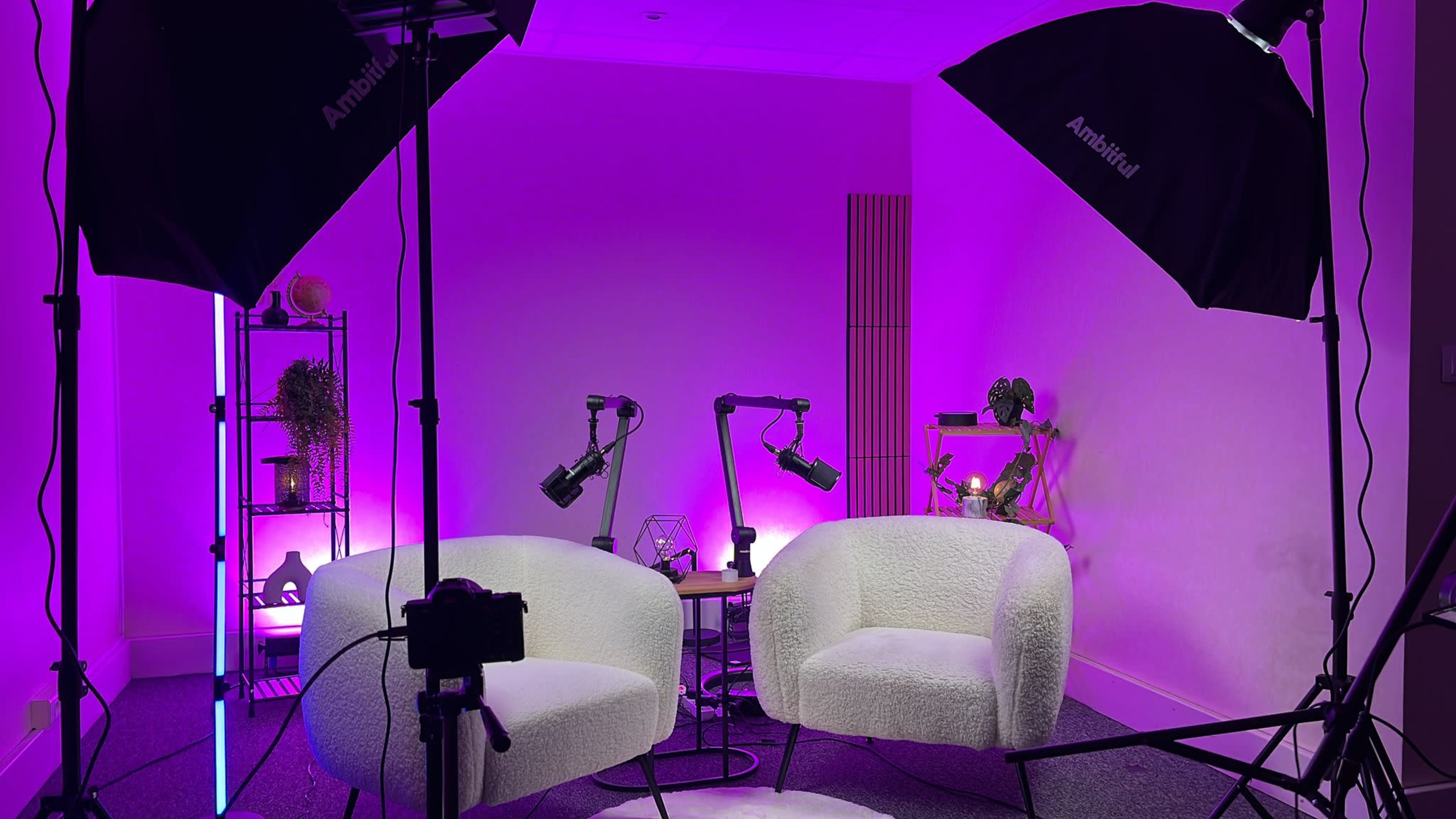 The image shows a modern studio setup with two white chairs, lighting equipment, and décor featuring purple accents.