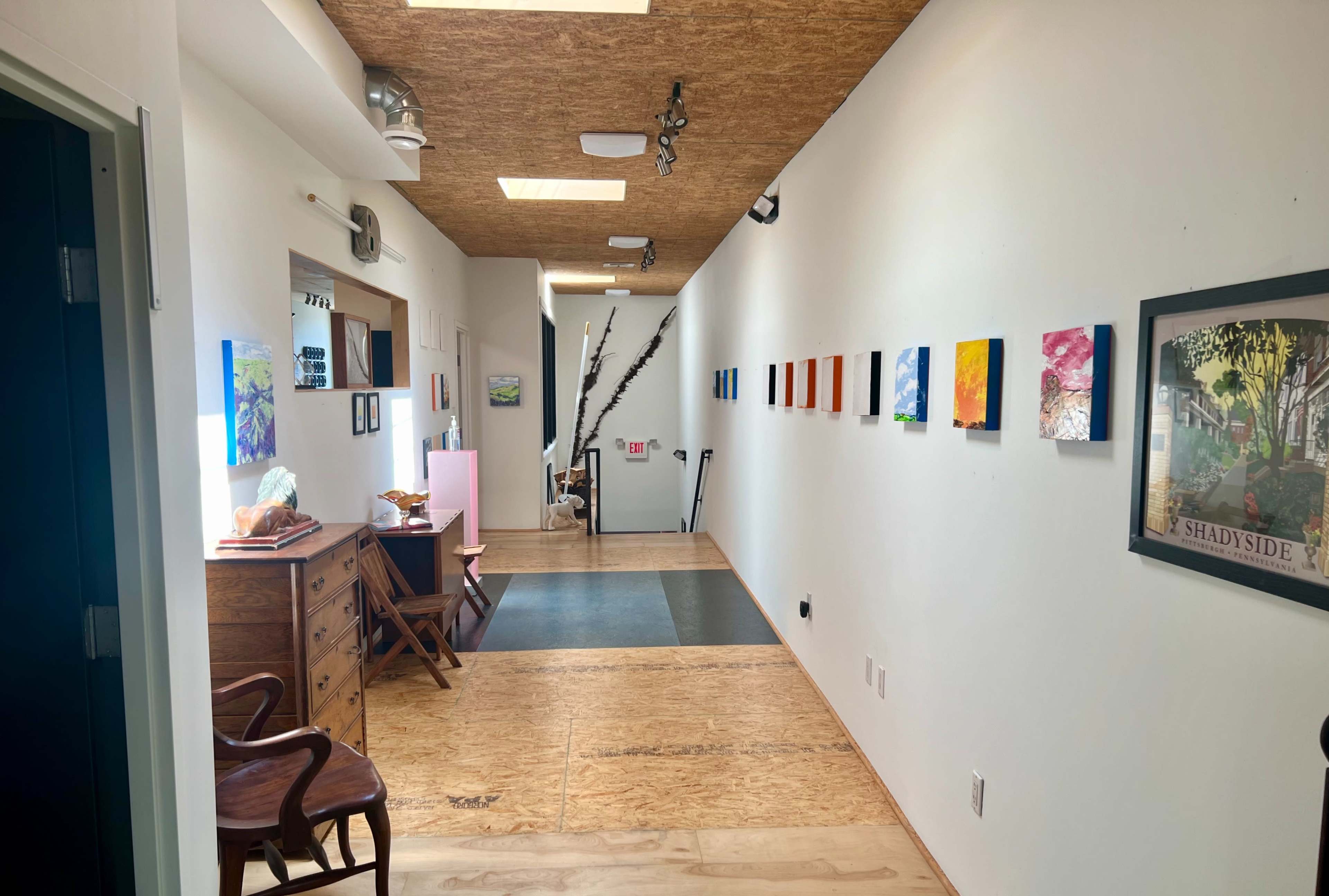 A hallway with wooden flooring, illuminated by skylights, and featuring colorful artwork on the walls alongside furniture and a staircase in the background.