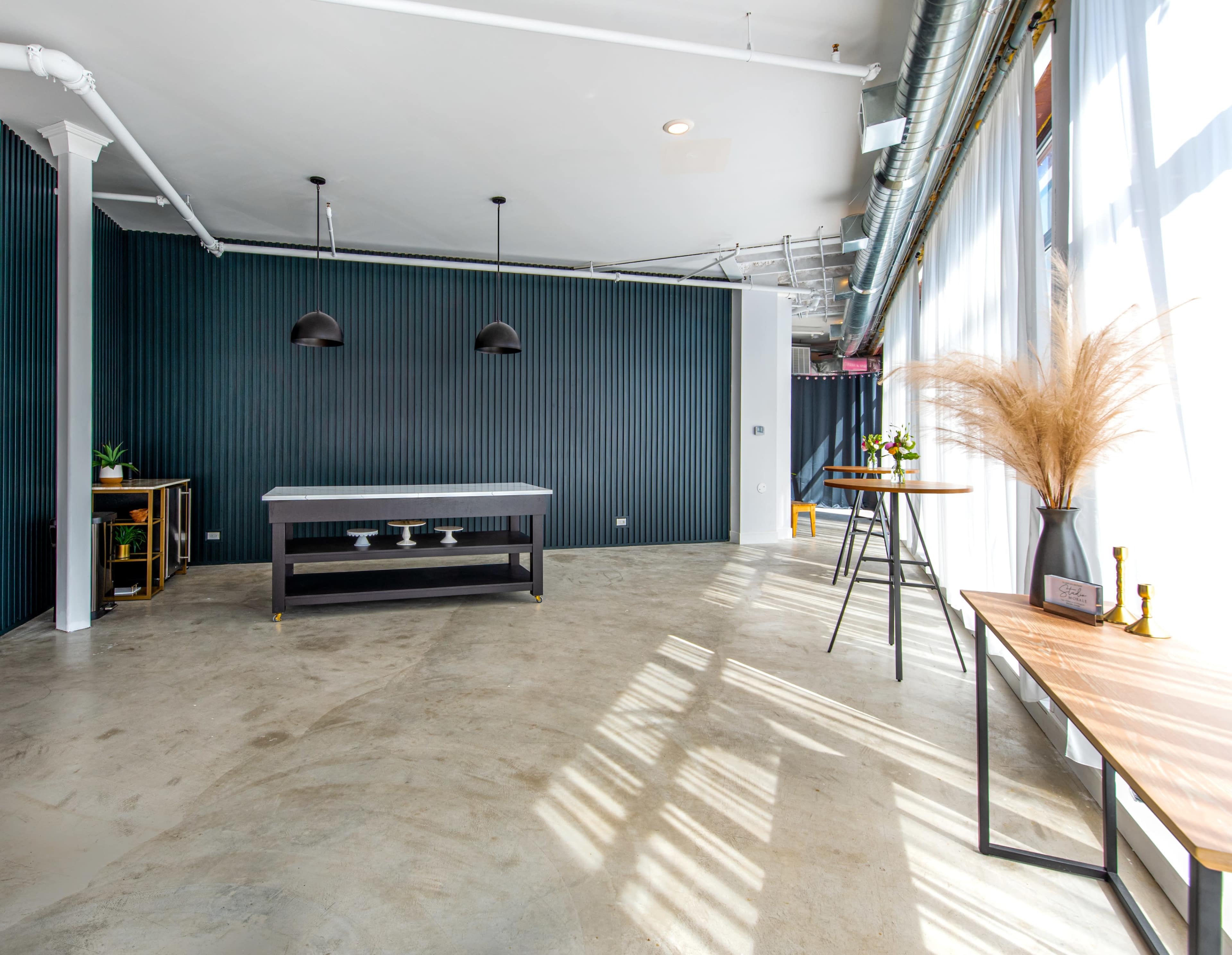 A modern, minimalistic interior space with a table in the center, black wall accents, and large windows allowing natural light to enter.