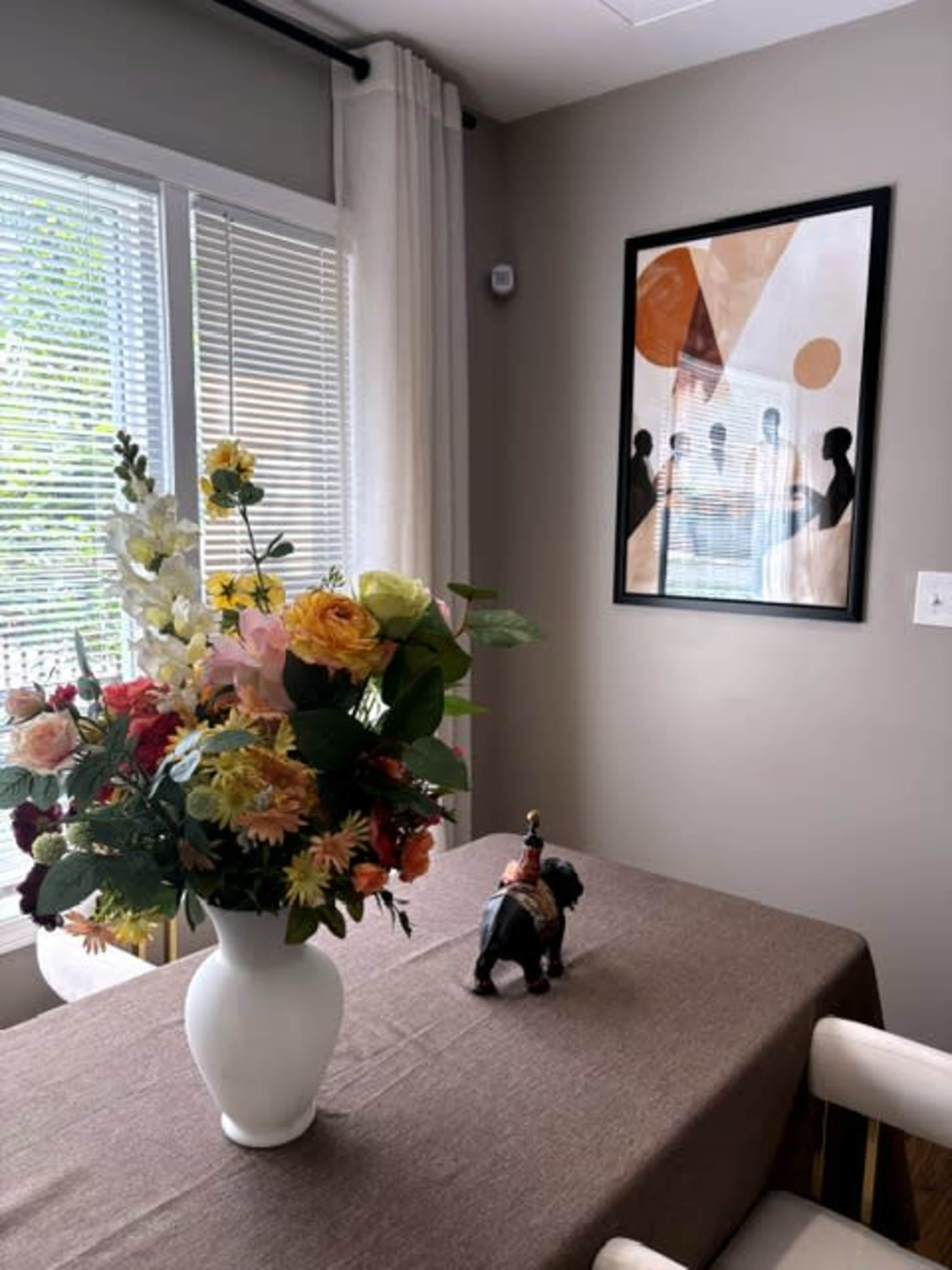 The image shows a dining table adorned with a large floral arrangement in a white vase, accompanied by a decorative figurine, set against a wall with an abstract artwork.
