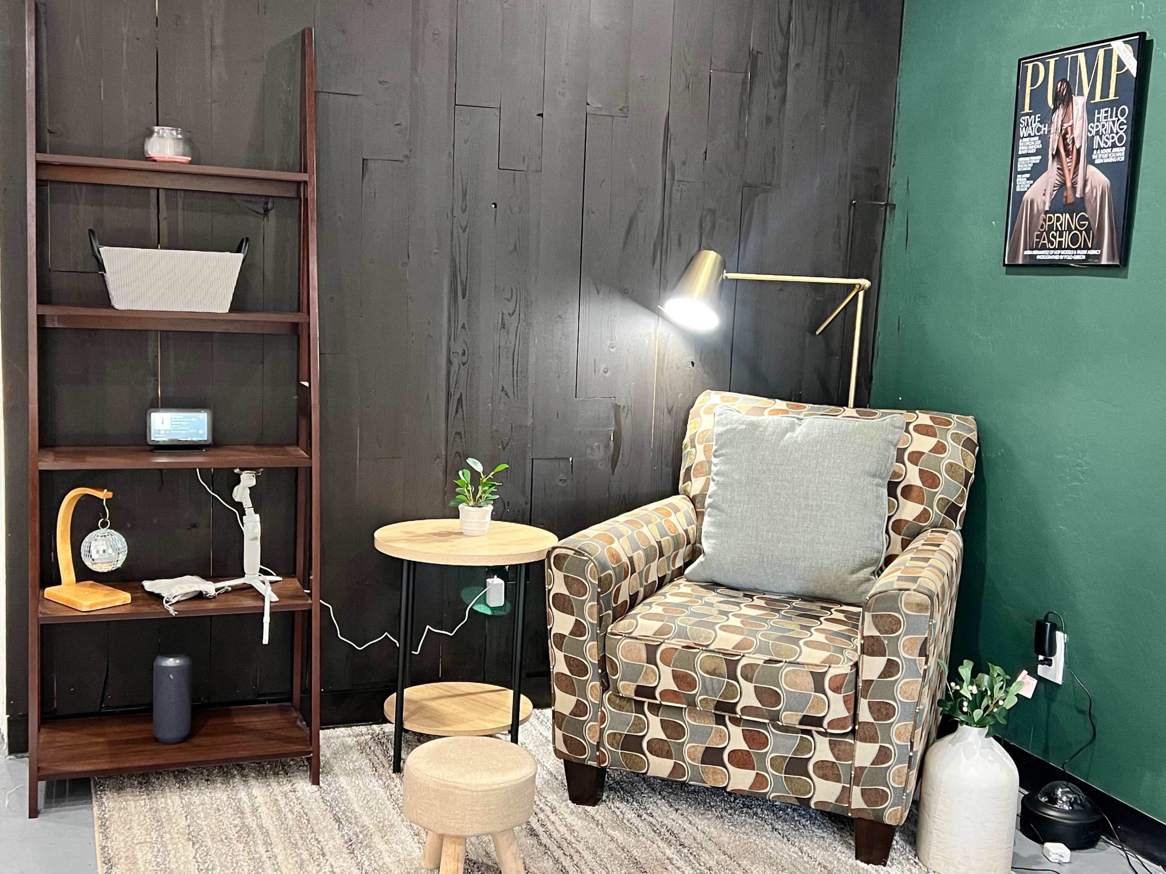A cozy living space with a patterned armchair, a small side table, a bookshelf with decor, and a floor lamp against a dark wood wall.