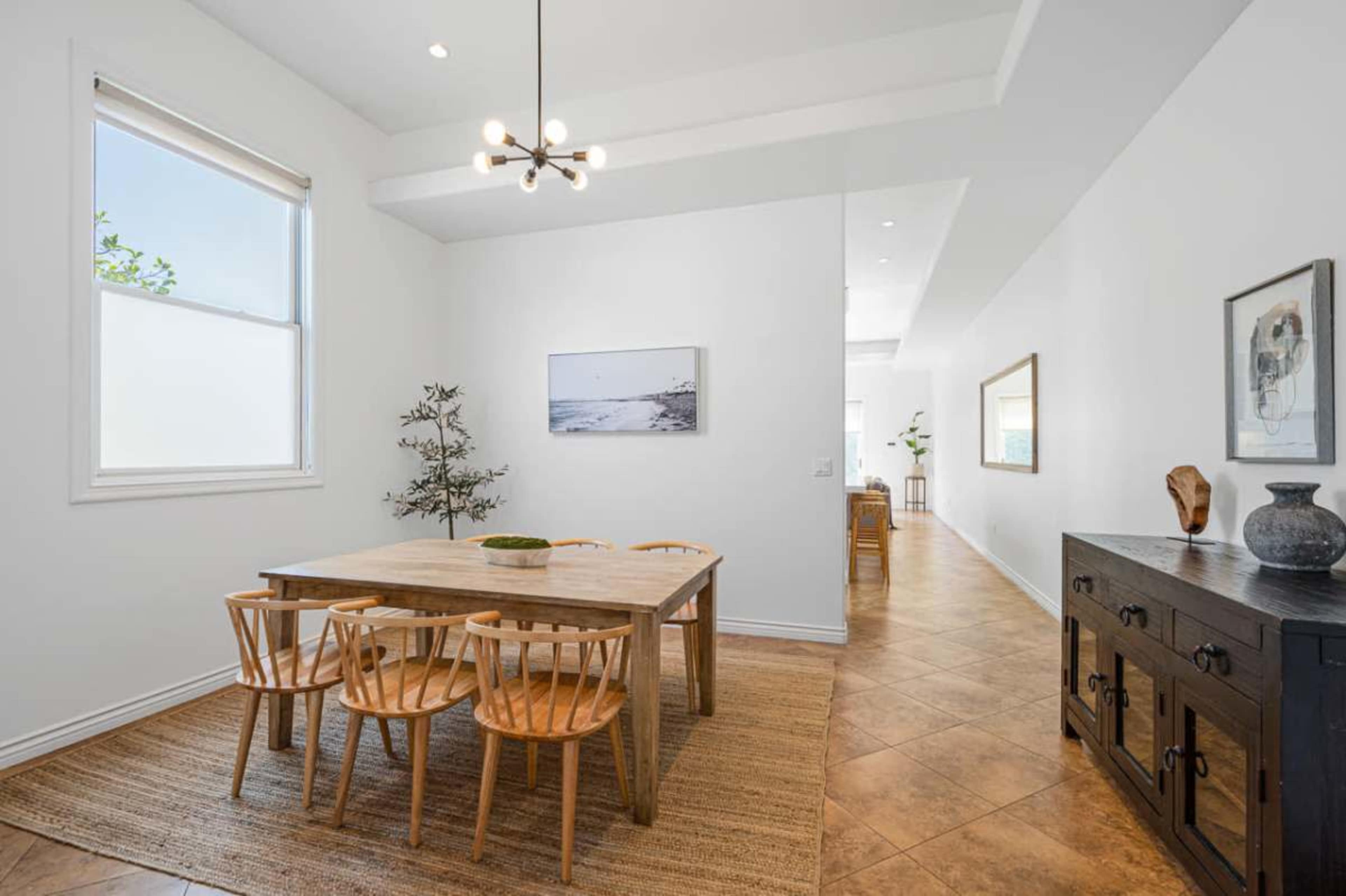 The image shows a bright dining area with a wooden table surrounded by six chairs, a decorative plant, and a hallway leading to another room.