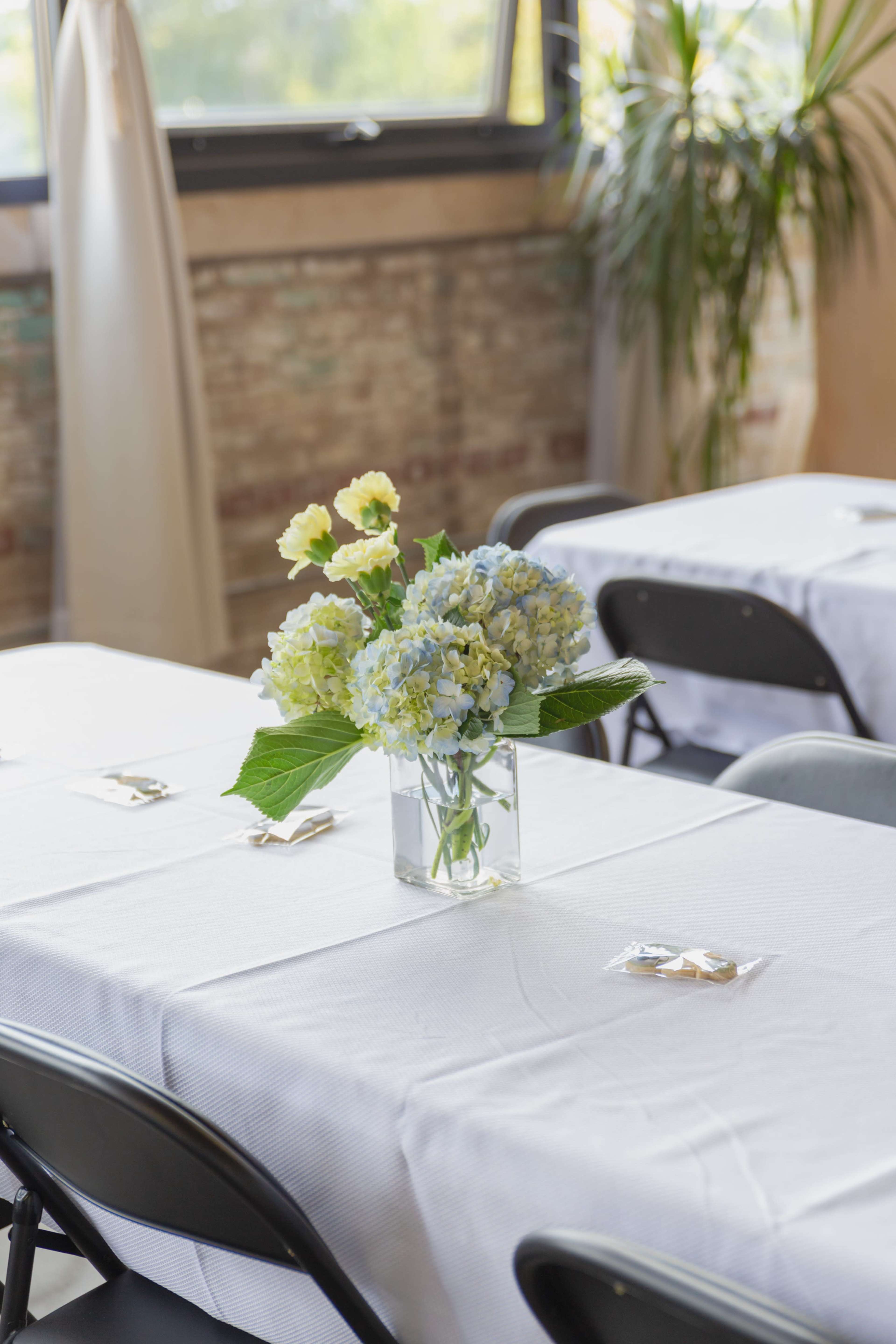 A clear vase filled with yellow and blue flowers sits on a white tablecloth-covered table, with black chairs arranged around it in a well-lit room.