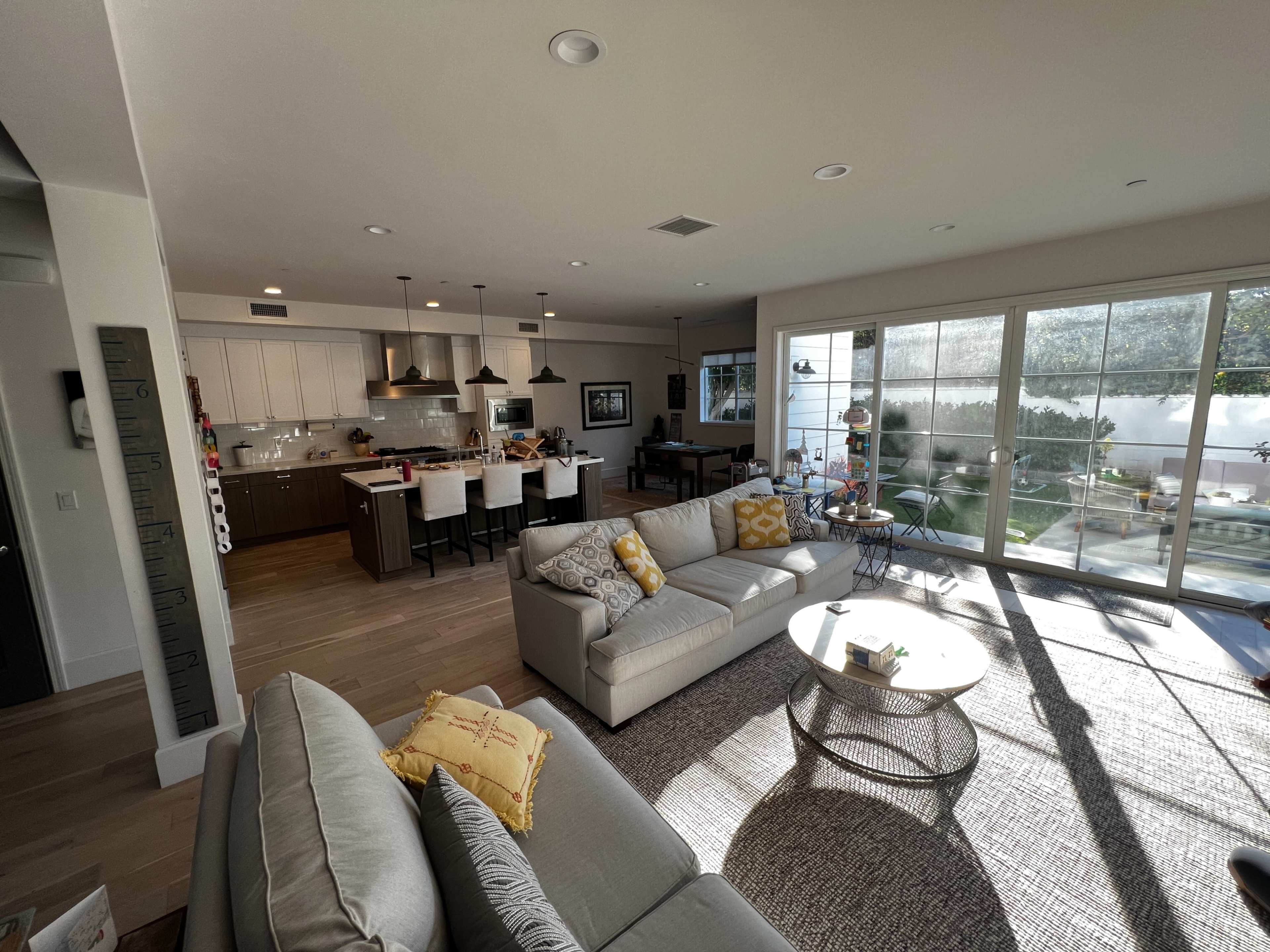 A modern living area features a light-colored sofa, a round coffee table, and an open kitchen with bar seating, all illuminated by natural light streaming through large windows.