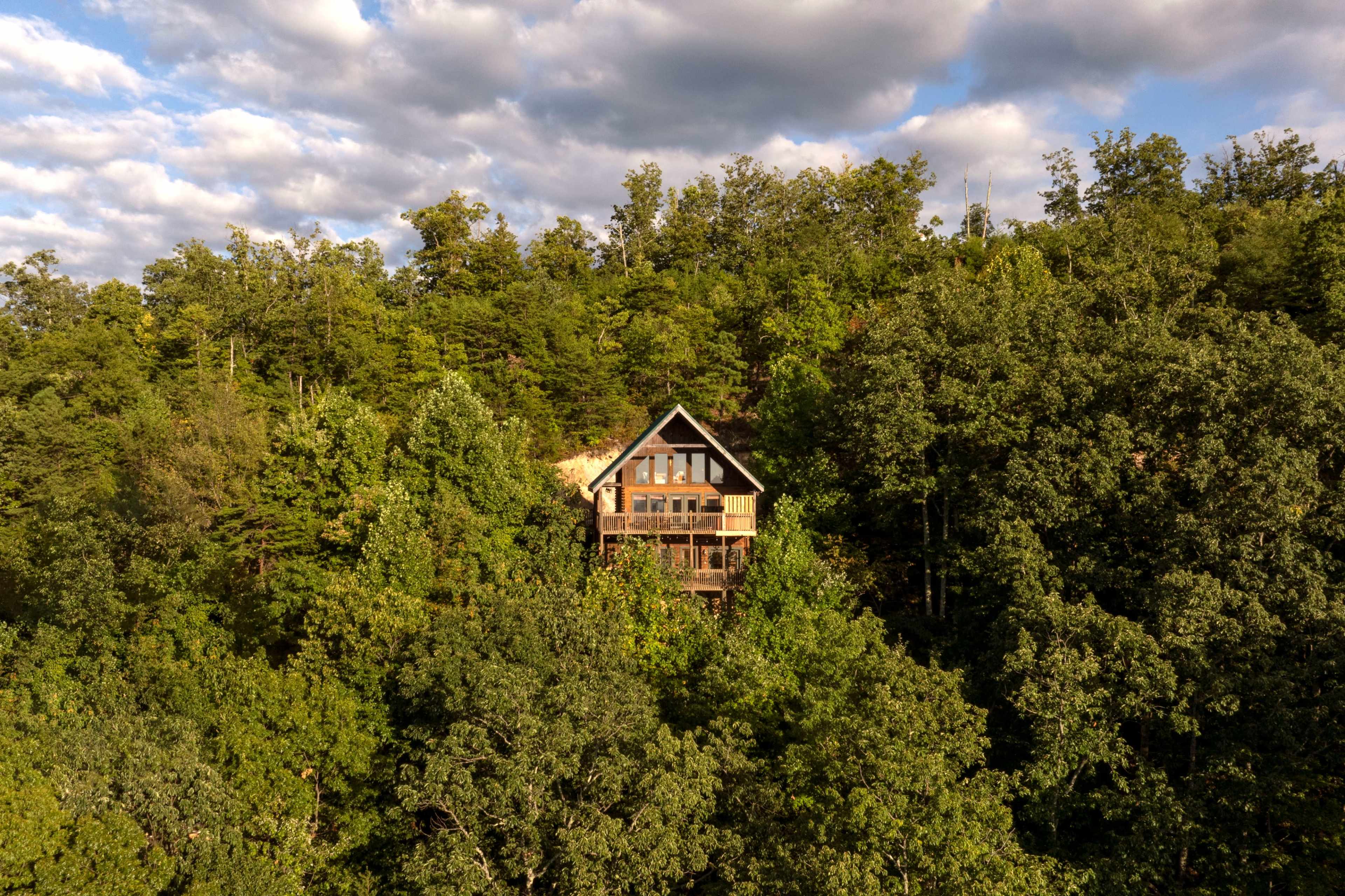 A large wooden cabin is nestled among dense green trees on a hillside under a partly cloudy sky.